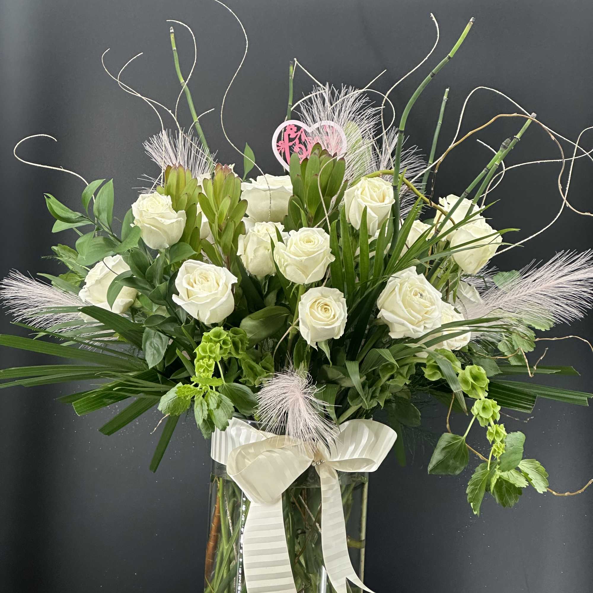 White roses arranged in a tall glass vase with greenery and a ribbon bow