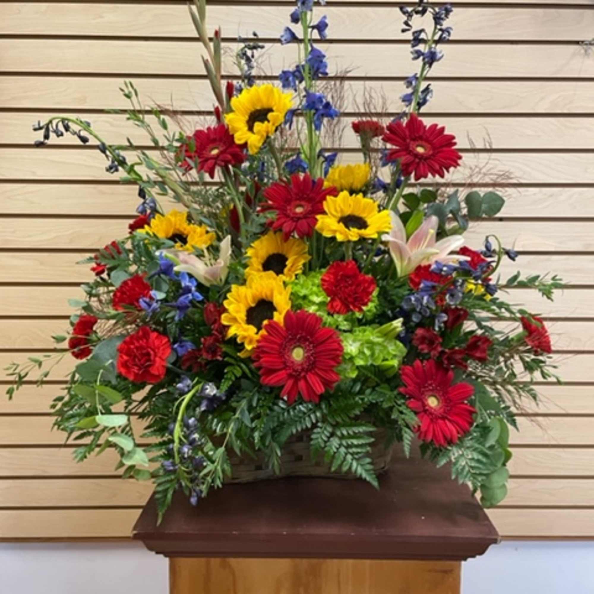 Large mixed flower arrangement with sunflowers, red gerberas, and blue delphinium in a basket