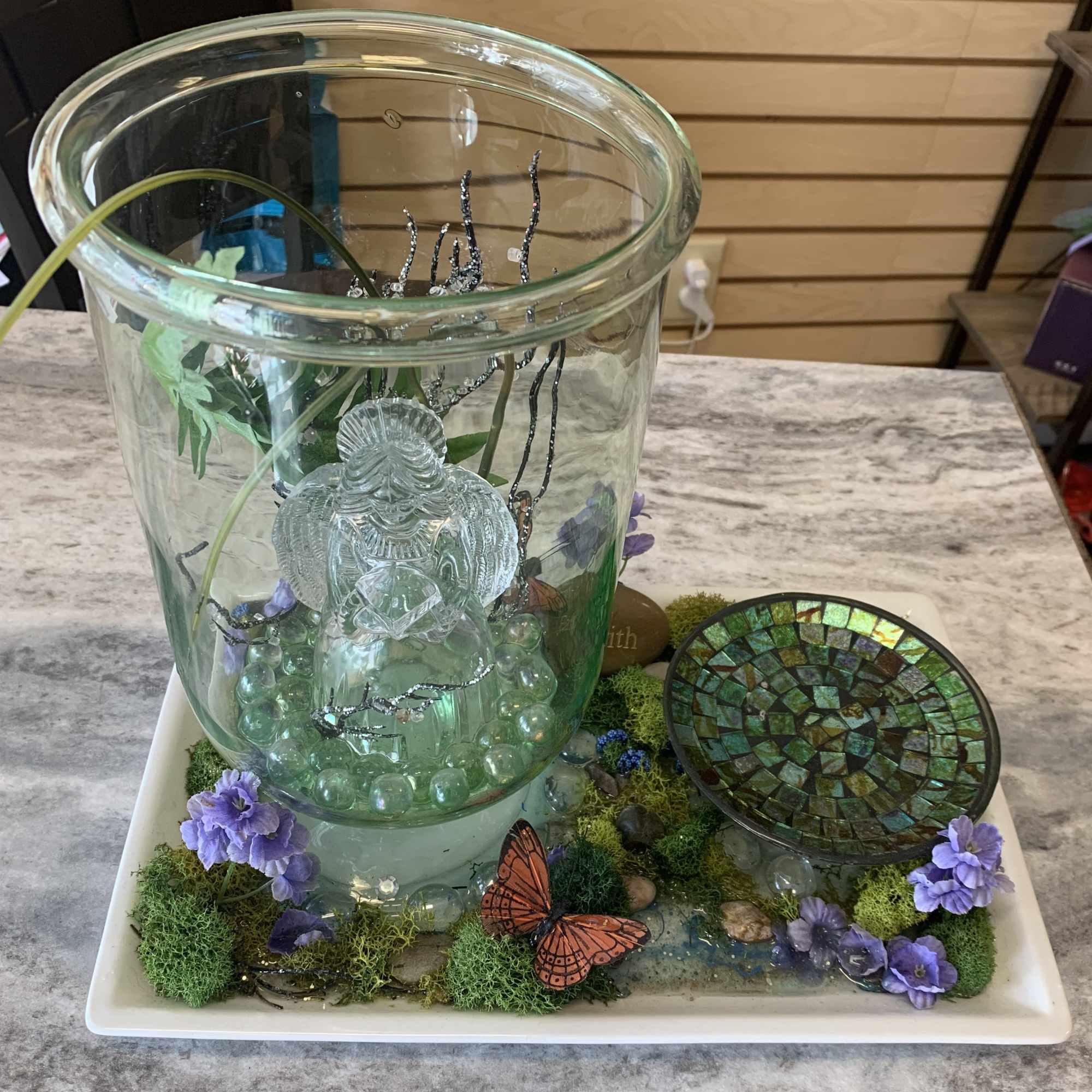 Glass fountain display with purple flowers and butterfly accents on a tray