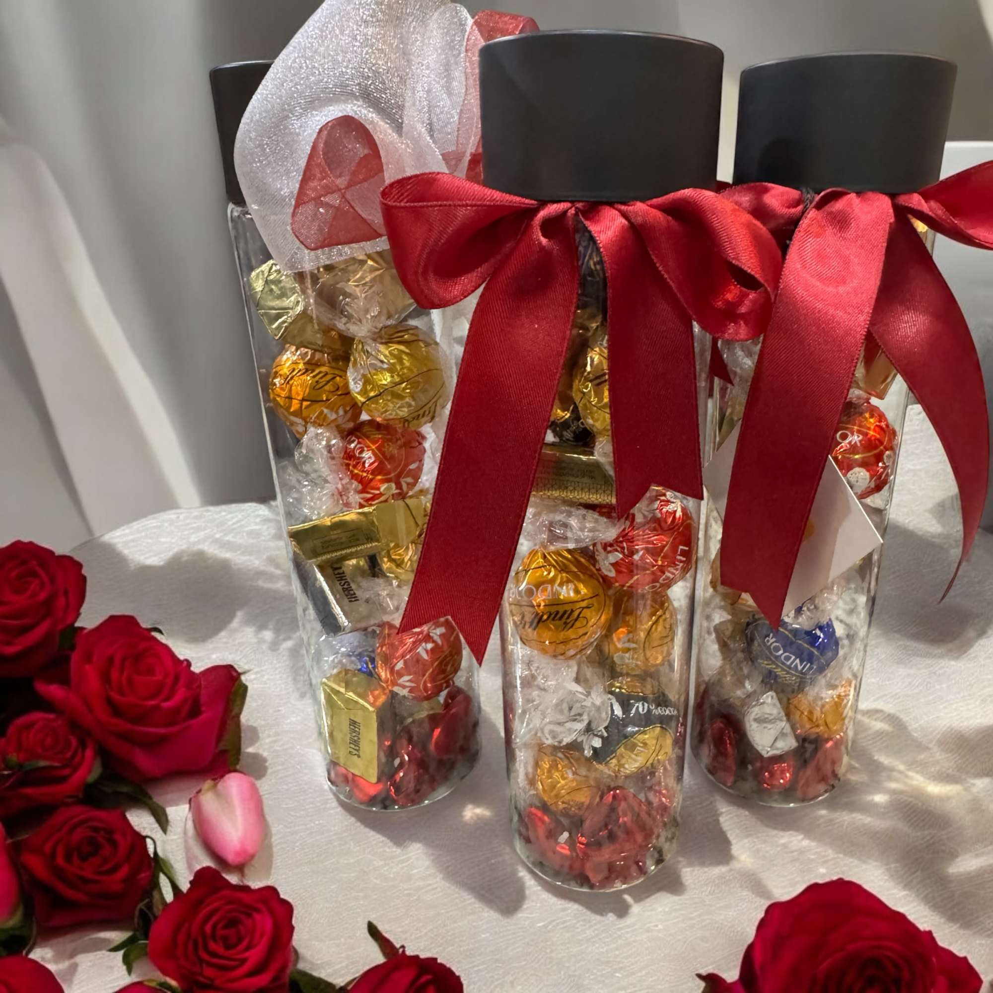 Clear gift tubes filled with chocolates and tied with red ribbons beside red roses