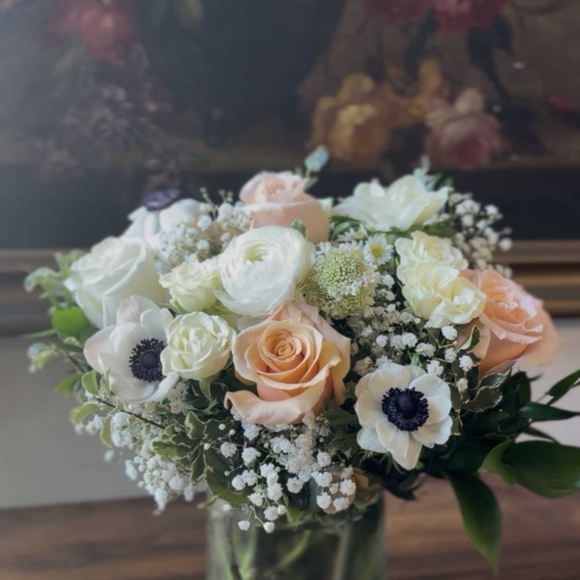 Round arrangement of white and peach roses, ranunculus, and anemones in a clear glass vase.