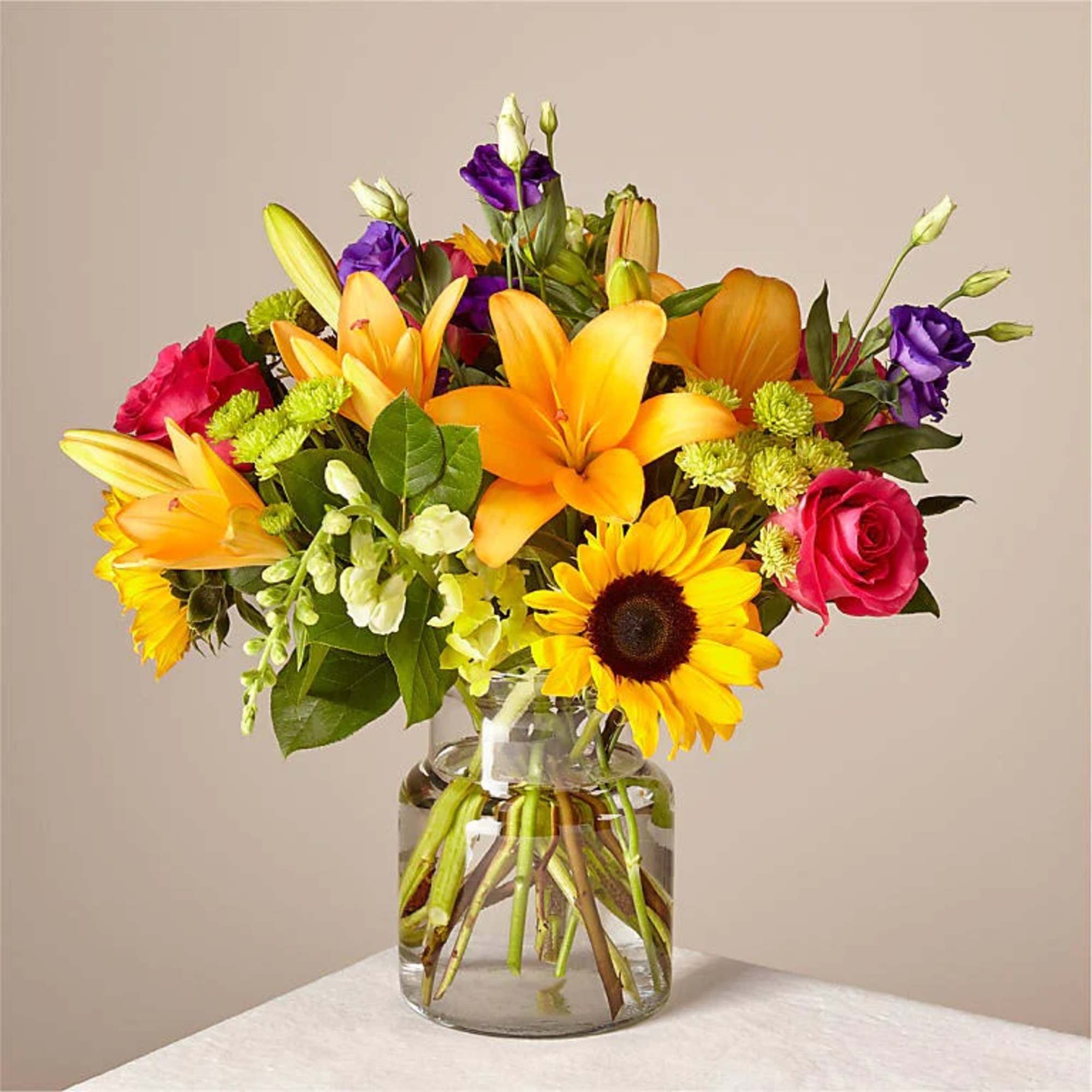 Mixed bouquet of lilies, roses, sunflowers, and purple blooms in a glass vase