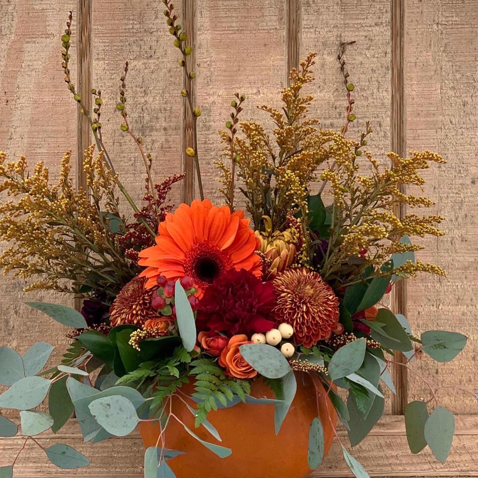 Autumn arrangement of orange gerbera, roses, and mums in a pumpkin container with eucalyptus accents