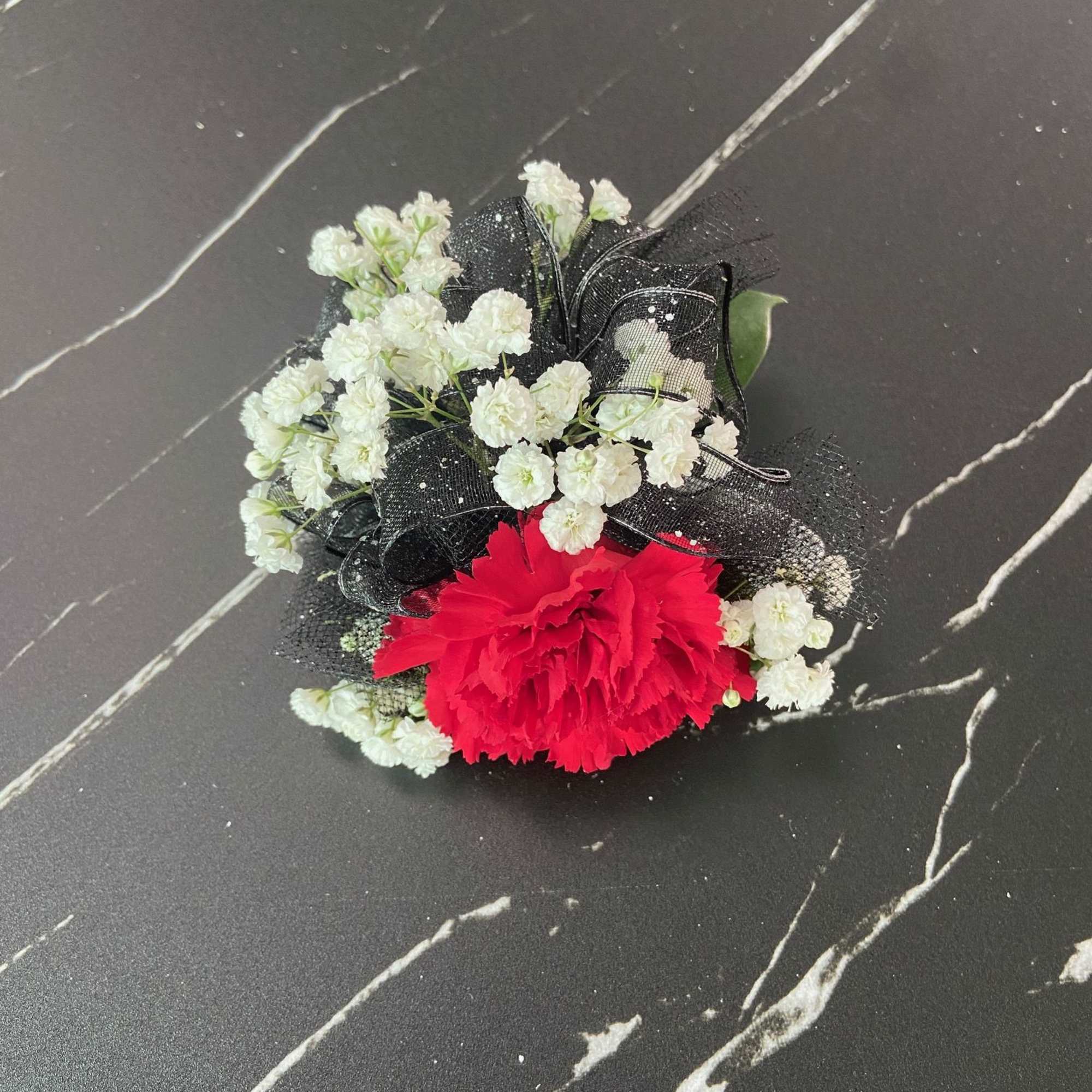 Red carnation corsage with white filler flowers and black ribbon on a dark surface