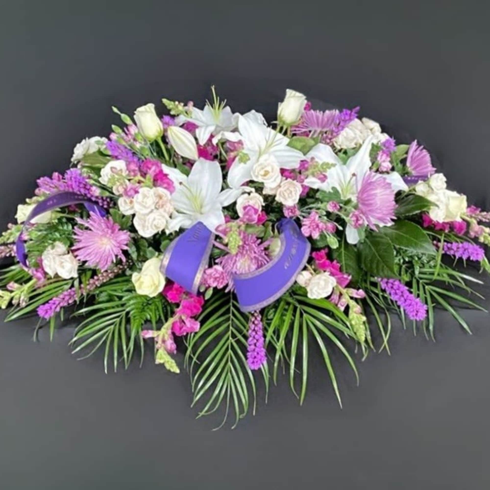 Elongated arrangement of white lilies, cream roses, and pink and purple flowers with palm leaves and purple ribbon
