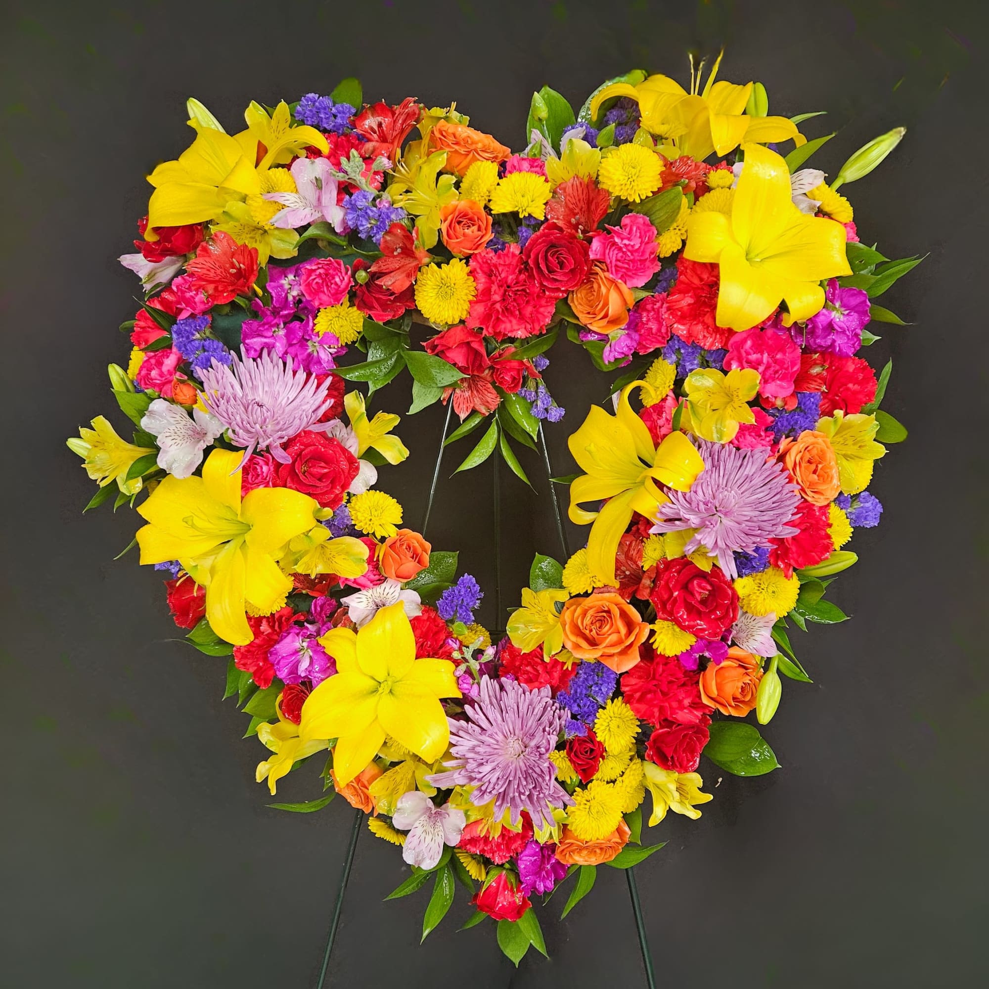 Heart-shaped standing wreath of bright yellow, pink, red, and purple flowers on a metal easel.