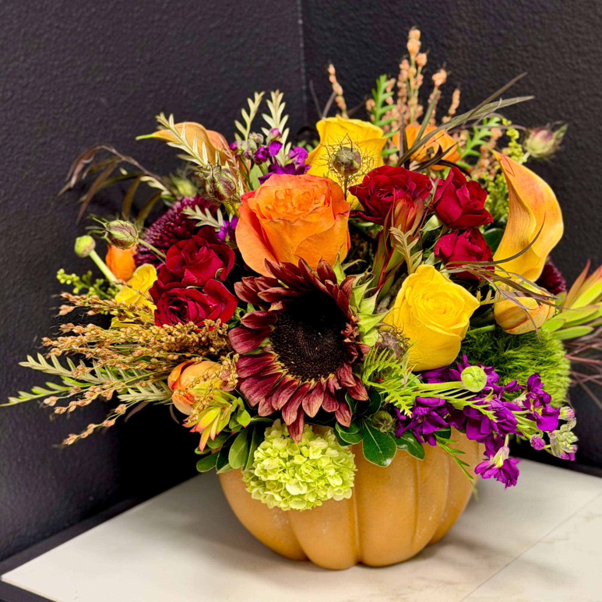 Mixed bouquet in a pumpkin-shaped container with roses, sunflowers, and calla lilies