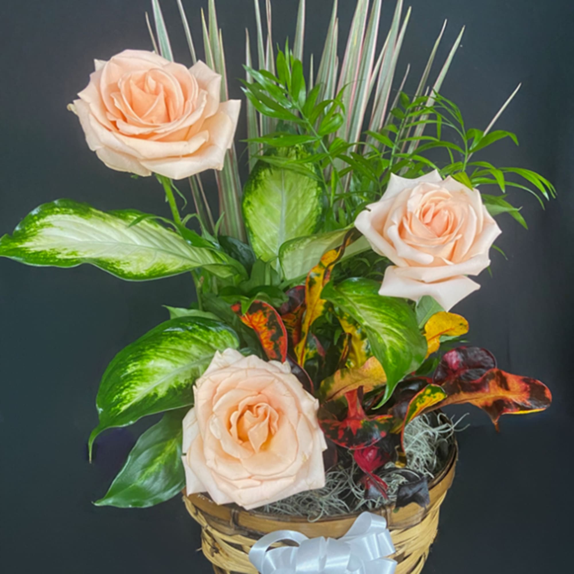 Peach roses arranged among mixed foliage plants in a woven basket with a white bow.