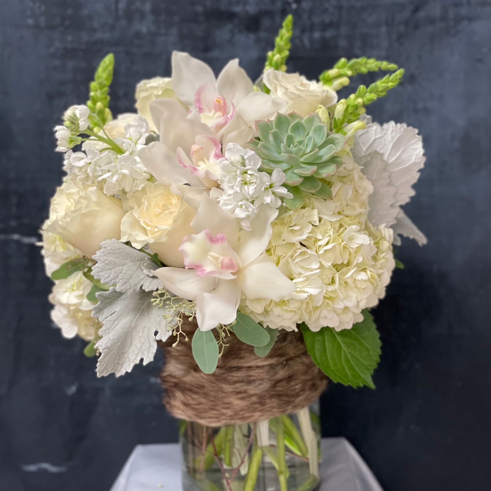 White hydrangeas, roses, orchids, and a succulent in a glass vase wrapped with brown twine