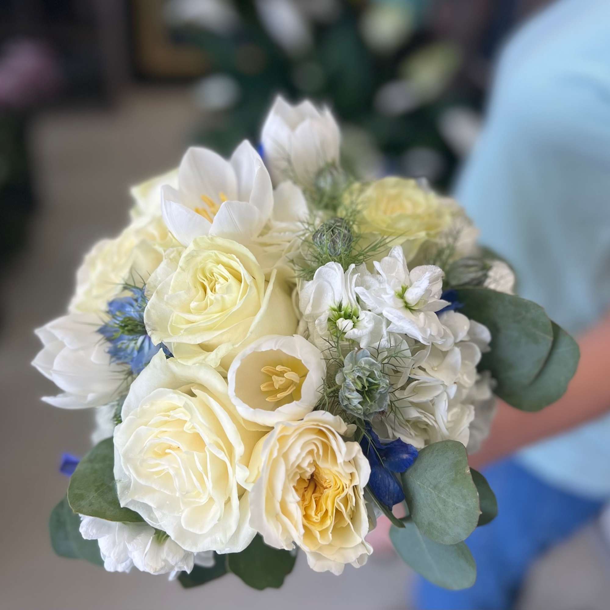 Handheld bouquet of ivory roses, white tulips, and soft blue accents in a compact round design