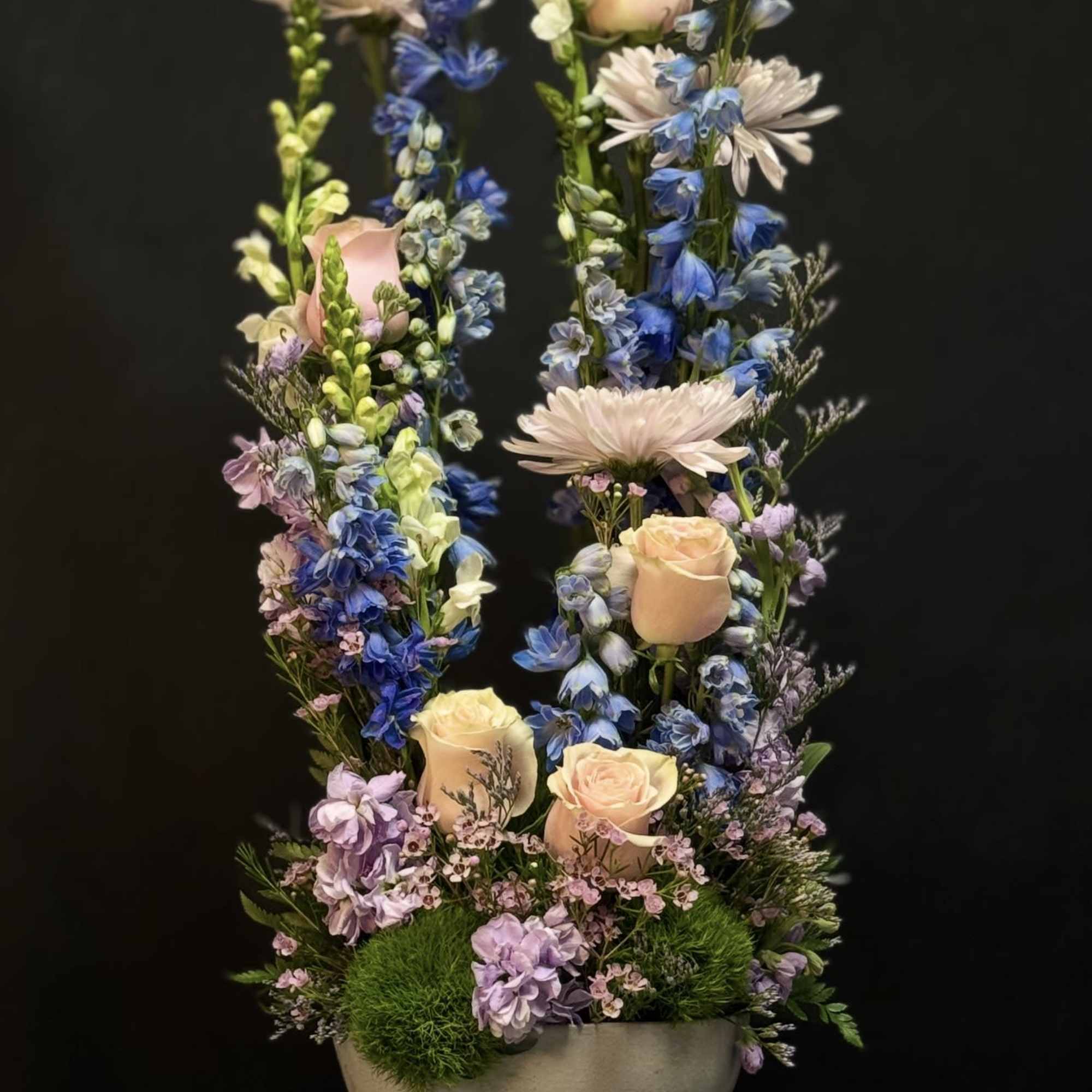 Tall pastel arrangement of peach roses and blue delphinium in a white bowl vase on draped fabric