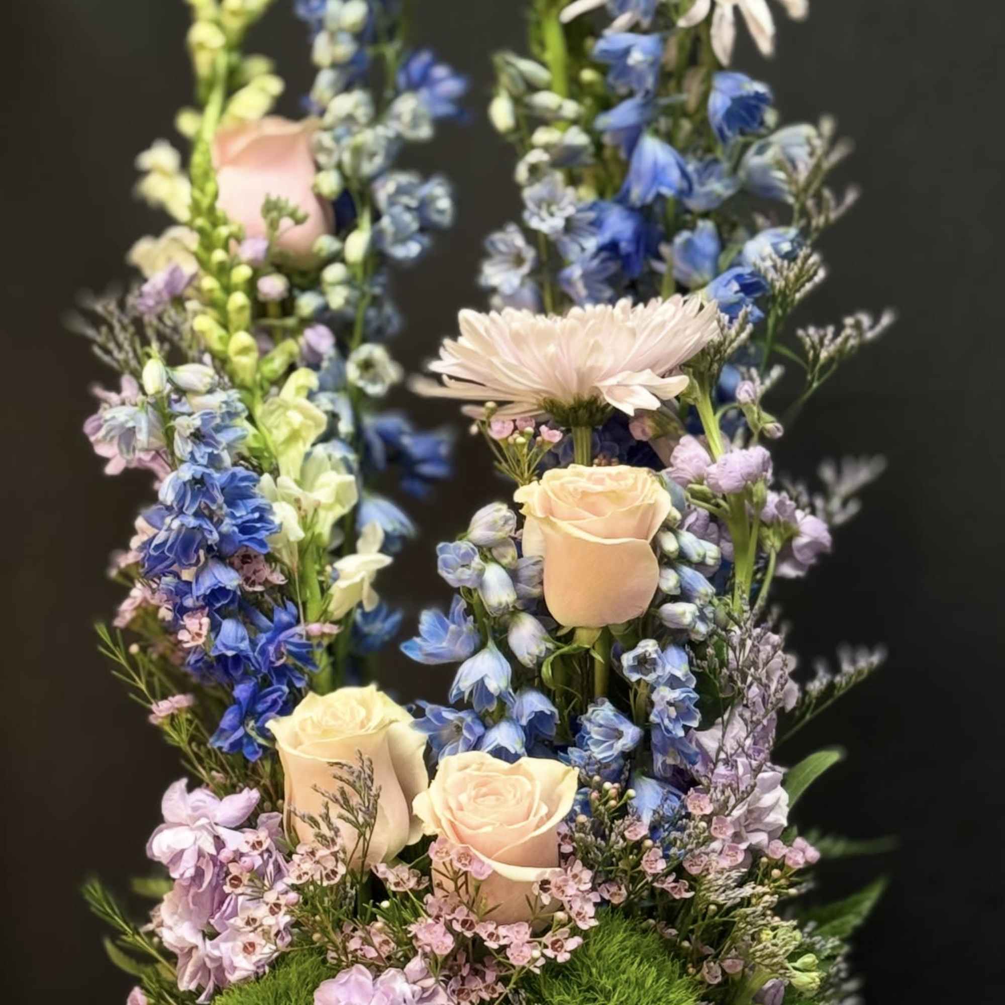 Tall pastel arrangement with peach roses, blue delphinium, and mums in a white bowl vase