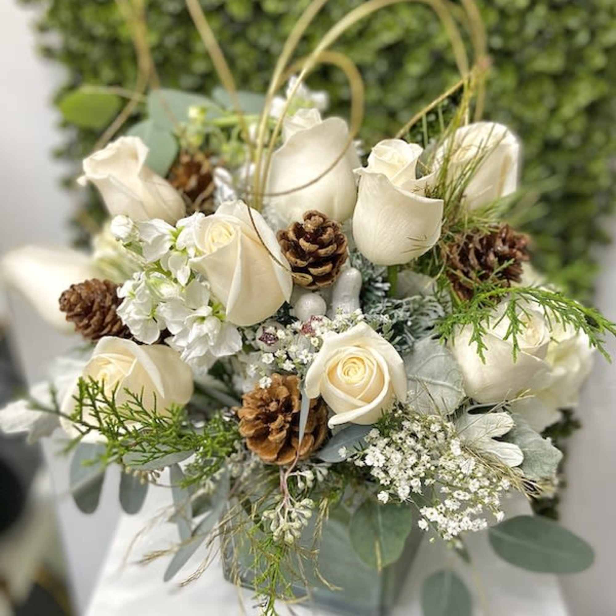 White rose arrangement with pinecones in a square vase against a green textured backdrop