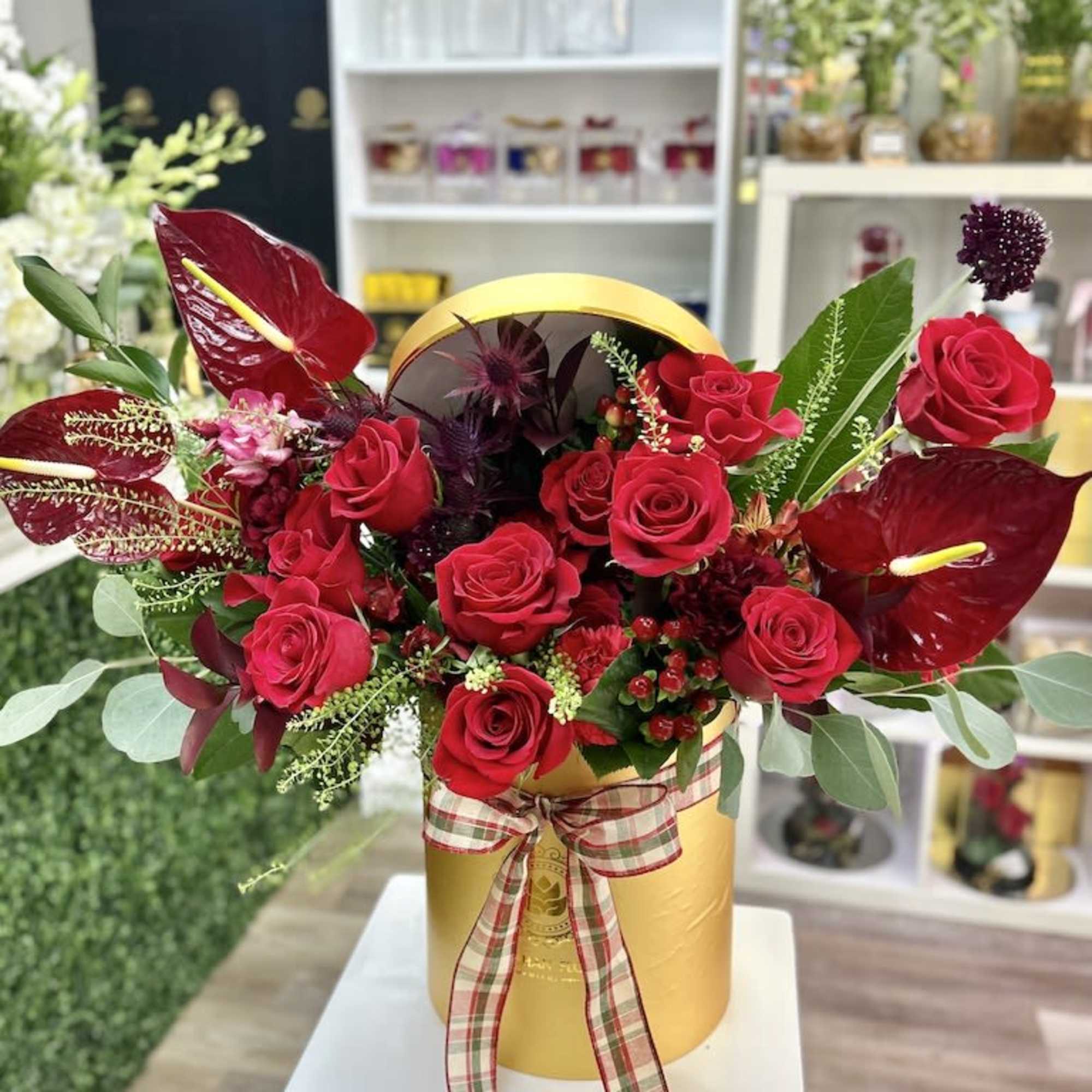 Red roses and dark red tropical blooms arranged in a gold hatbox with a plaid ribbon bow.