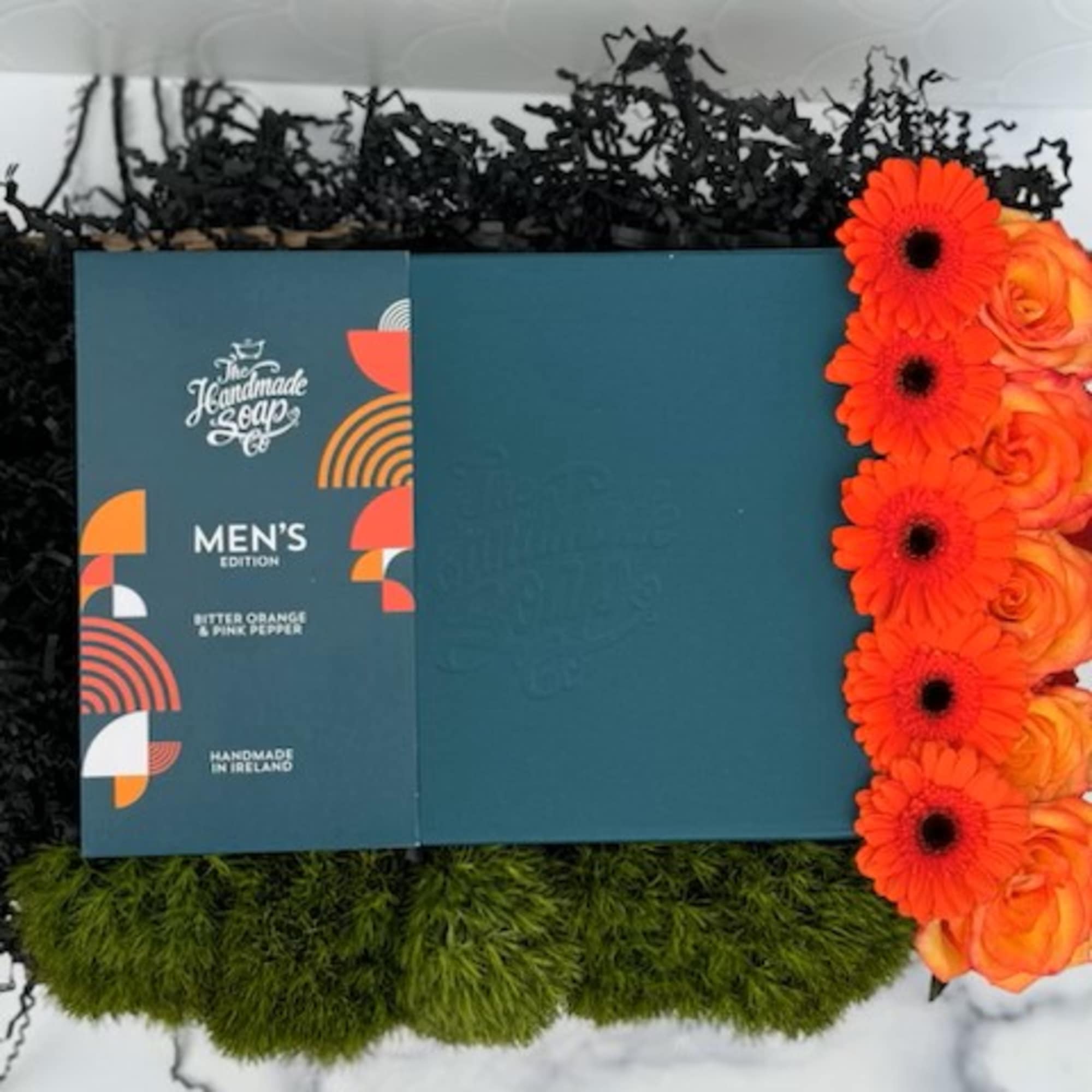 Men's soap gift box with bright orange gerbera daisies and roses, green moss and black filler in a tray