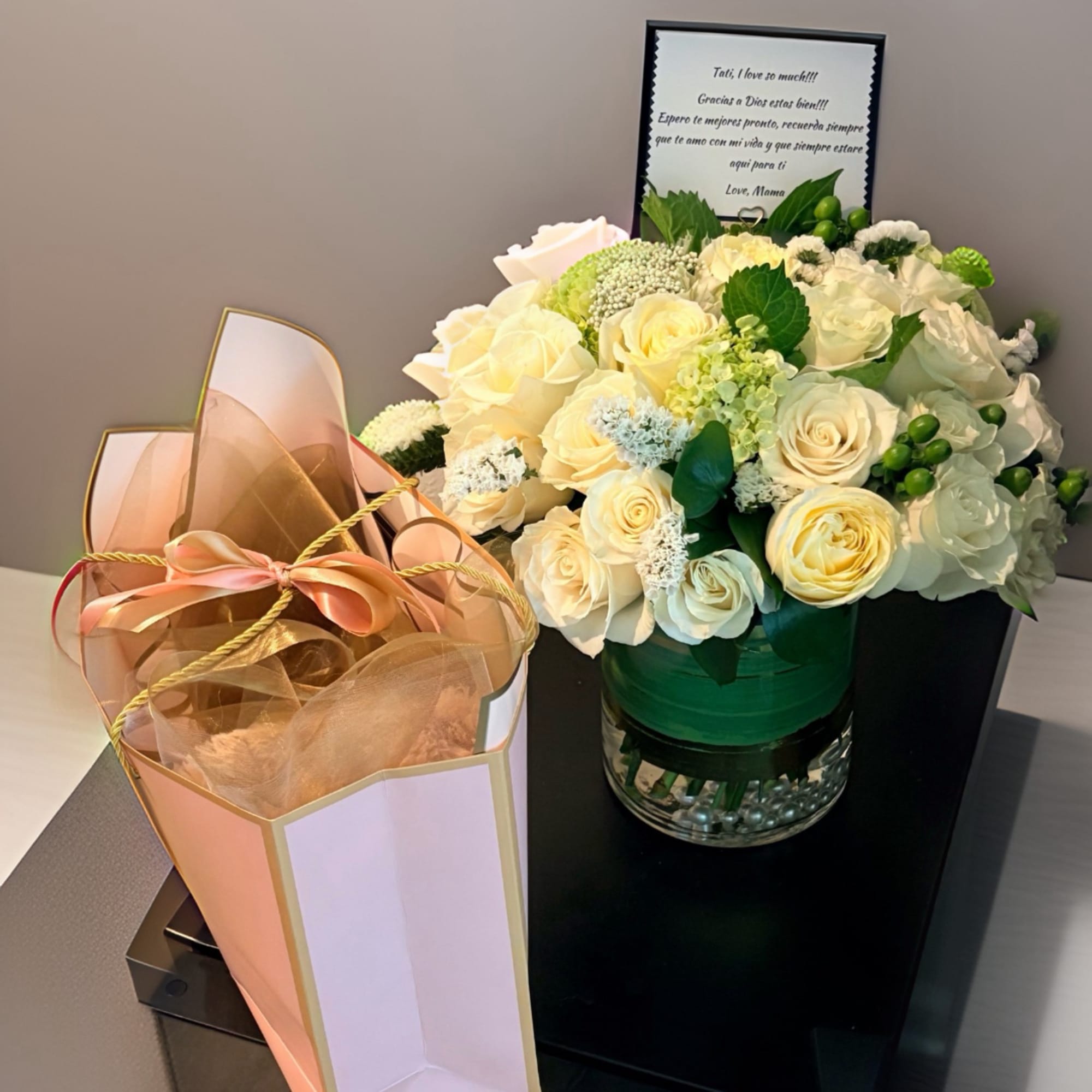 Cream rose arrangement in a glass vase beside a pink gift bag with ribbon.