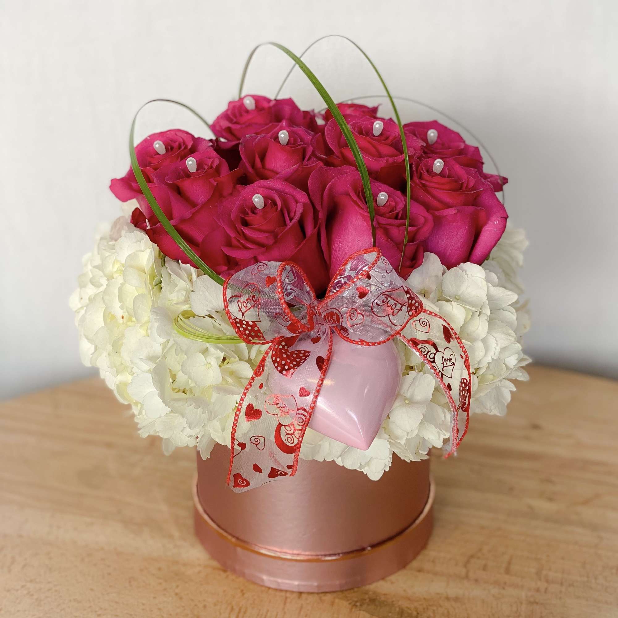 Pink roses arranged in a round box with white hydrangeas and a heart ribbon