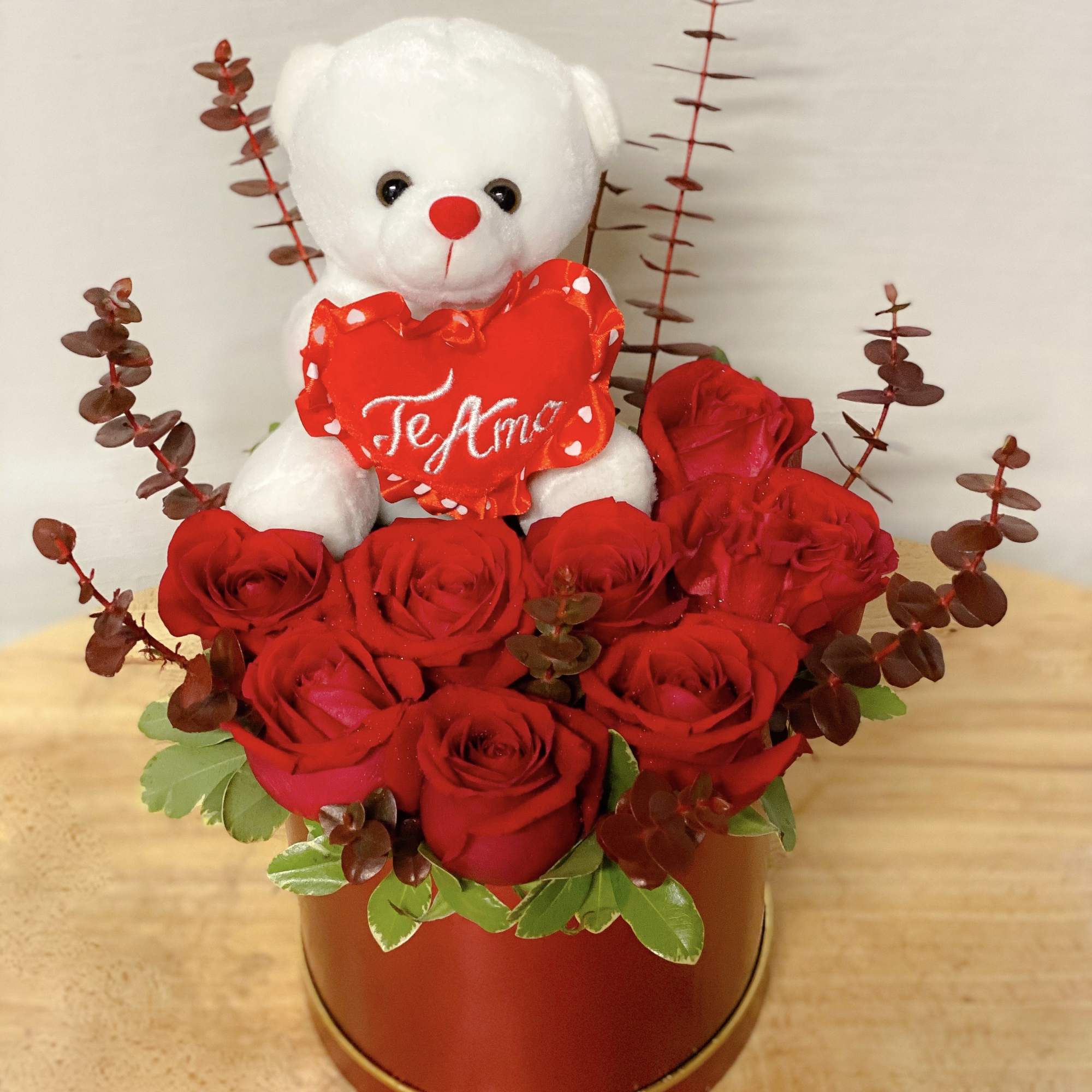 Red roses arranged with a white teddy bear holding a heart in a round box