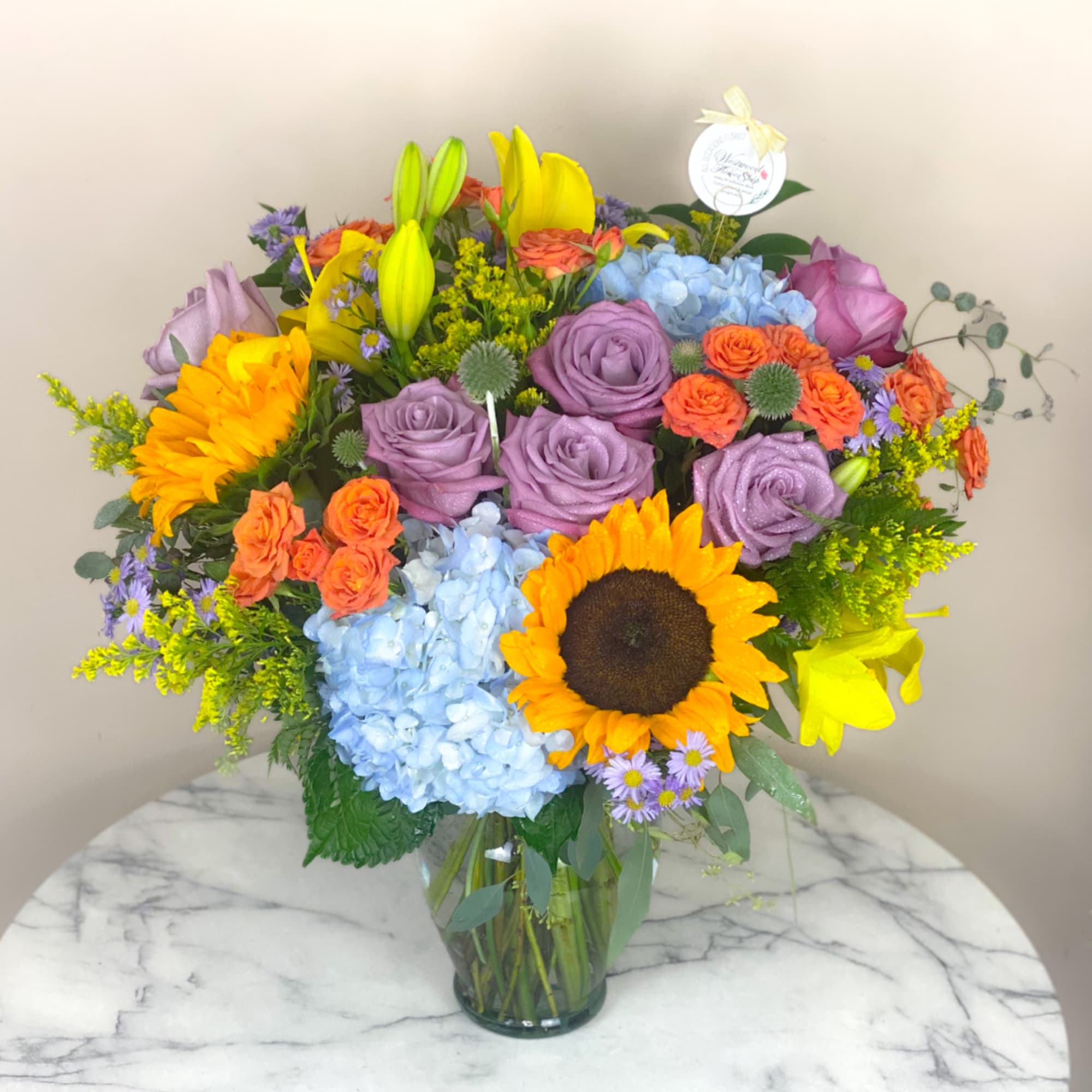 Colorful mixed bouquet in a clear glass vase with sunflowers, roses, lilies, and hydrangeas.