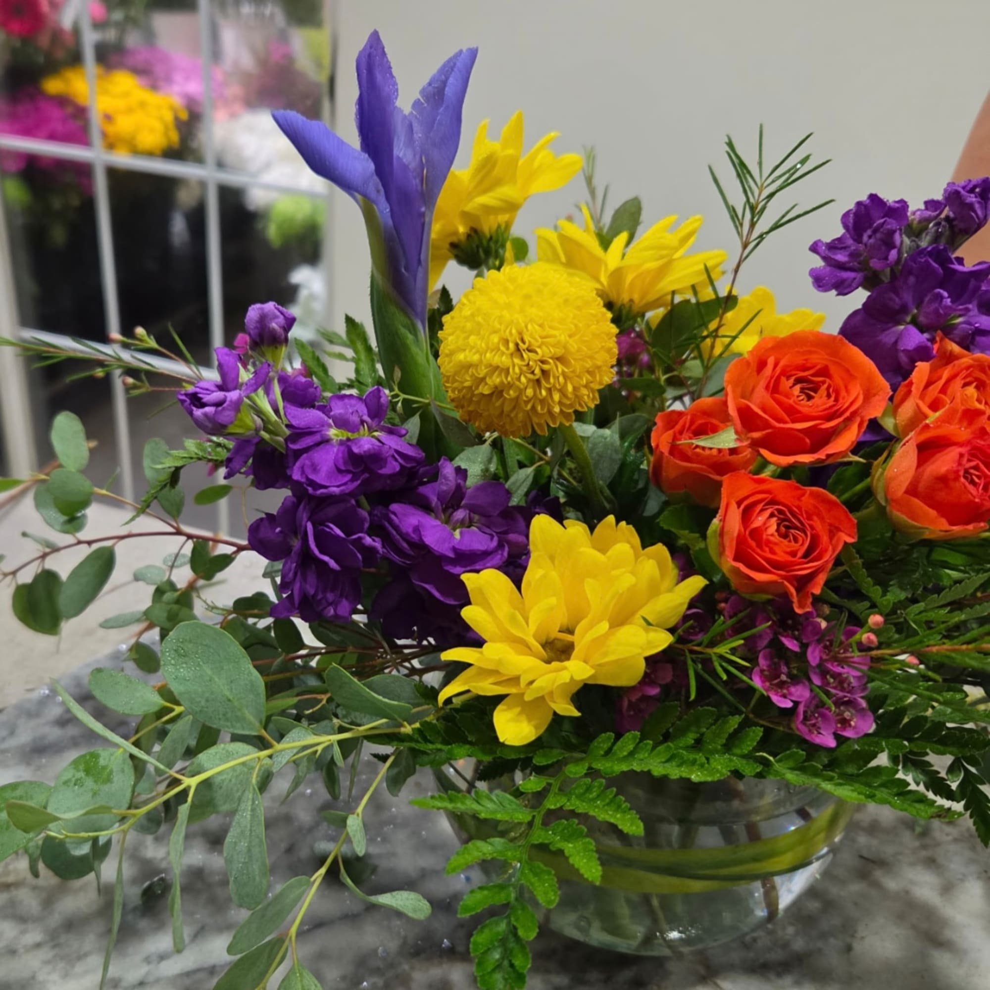 Colorful mixed bouquet with orange roses, yellow mums, and purple flowers in a glass bowl