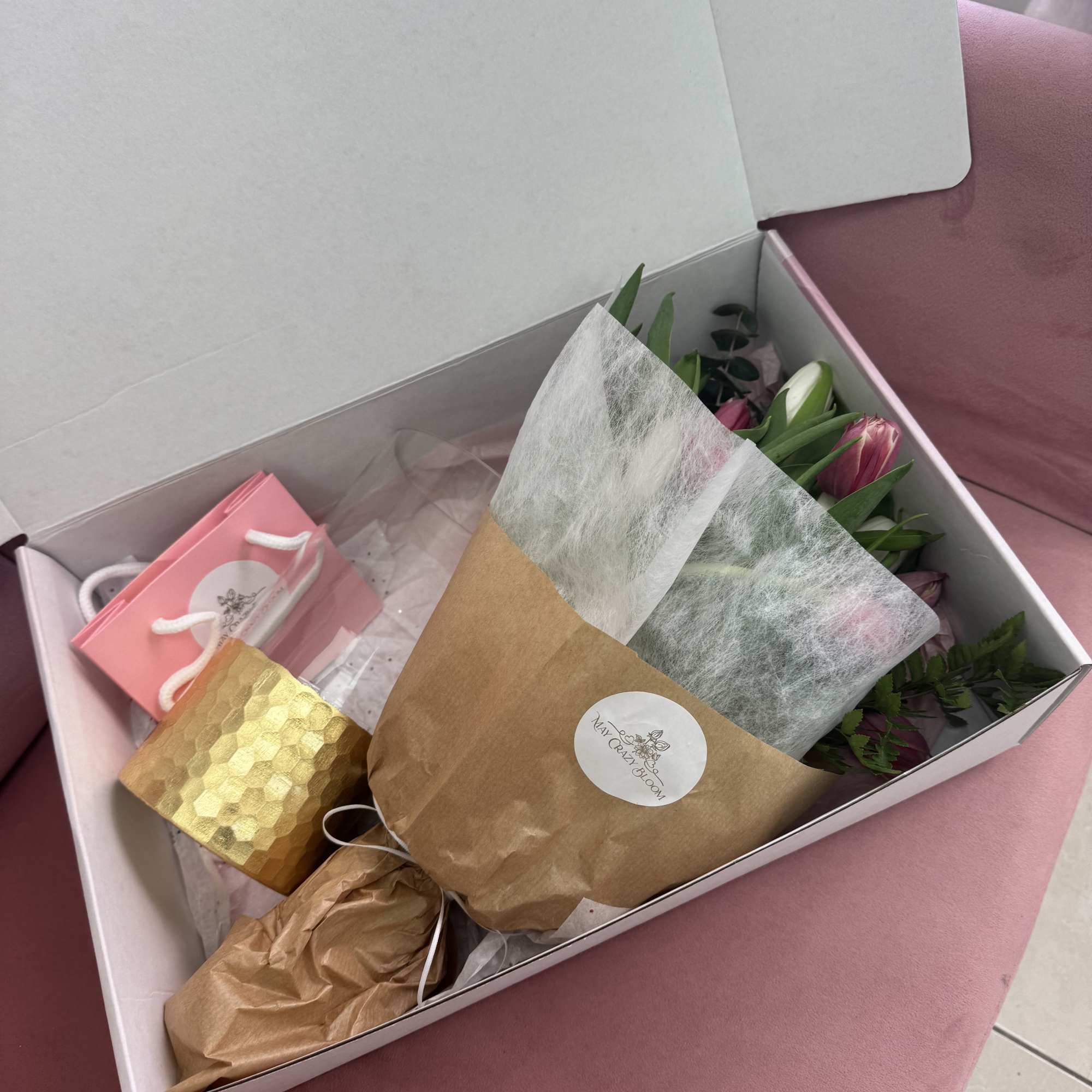 Pink tulips in a wrapped bouquet inside a gift box with a small pink bag.