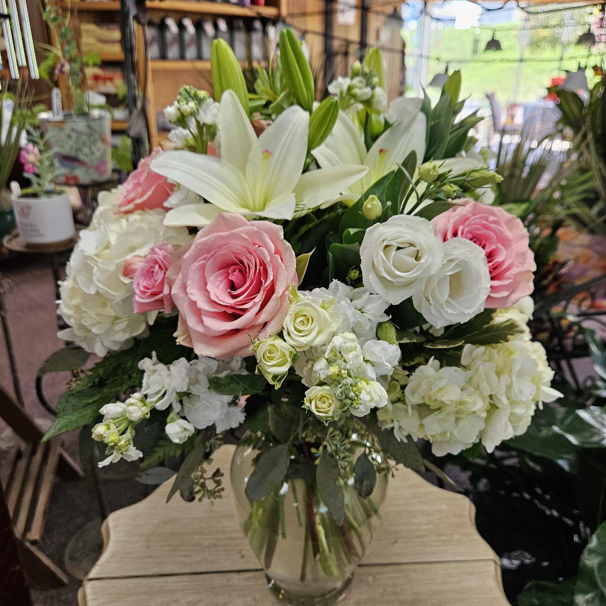 Pink roses, white lilies, and white hydrangeas arranged in a clear glass vase on a small wooden table