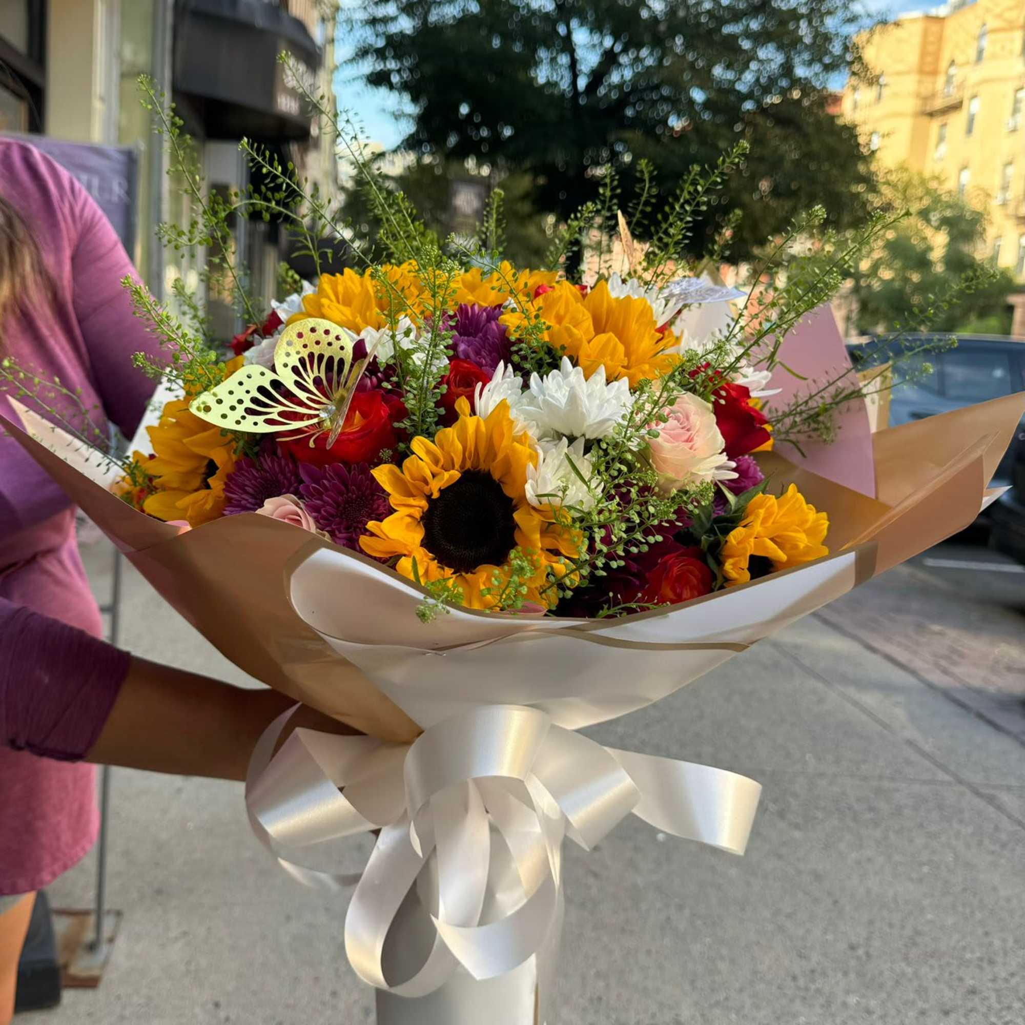 Handheld bouquet of sunflowers, roses, and daisies wrapped in cream paper with a ribbon