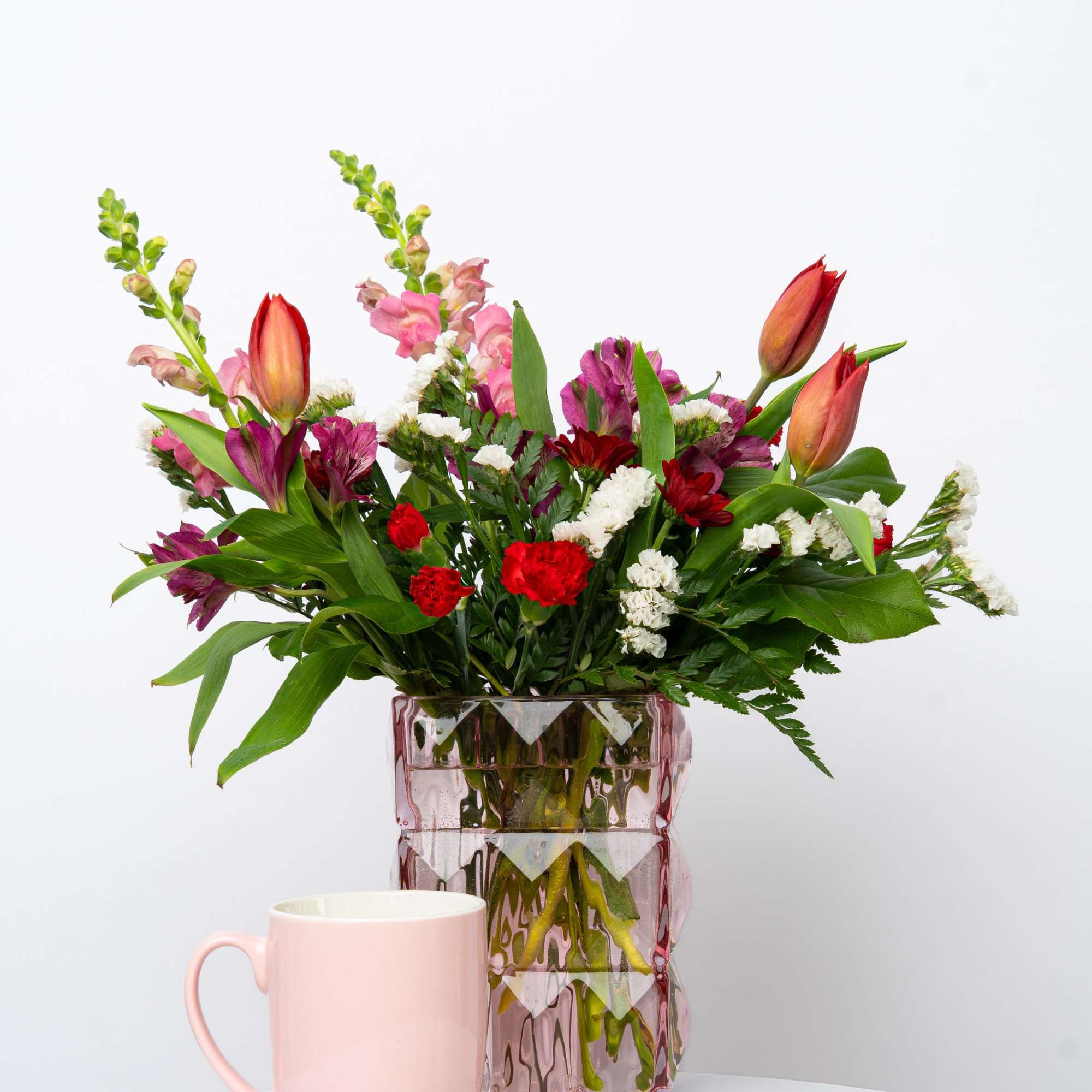Mixed bouquet in a pink glass vase with a pink mug in front