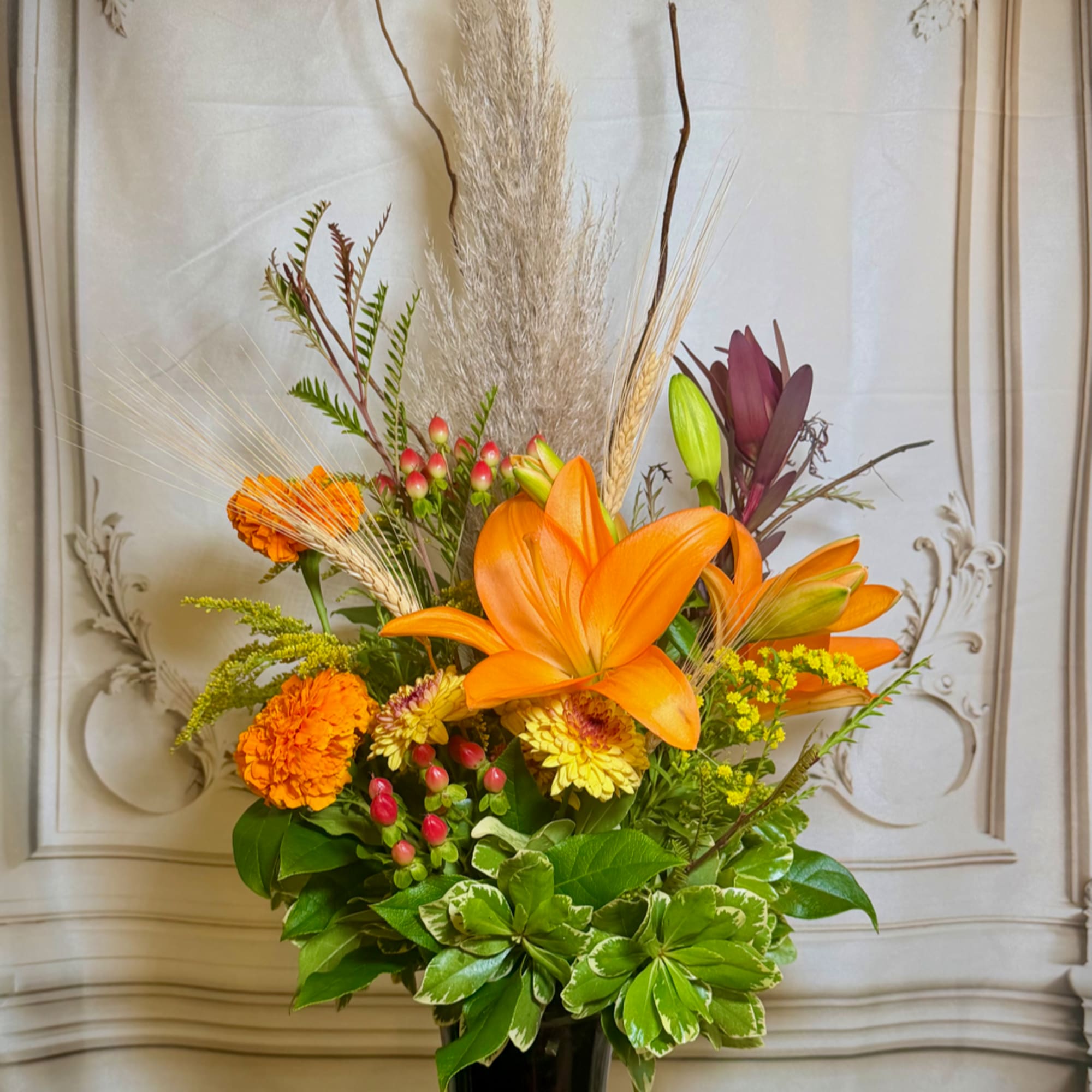 Orange lilies and marigolds in a tall vase with dried grasses