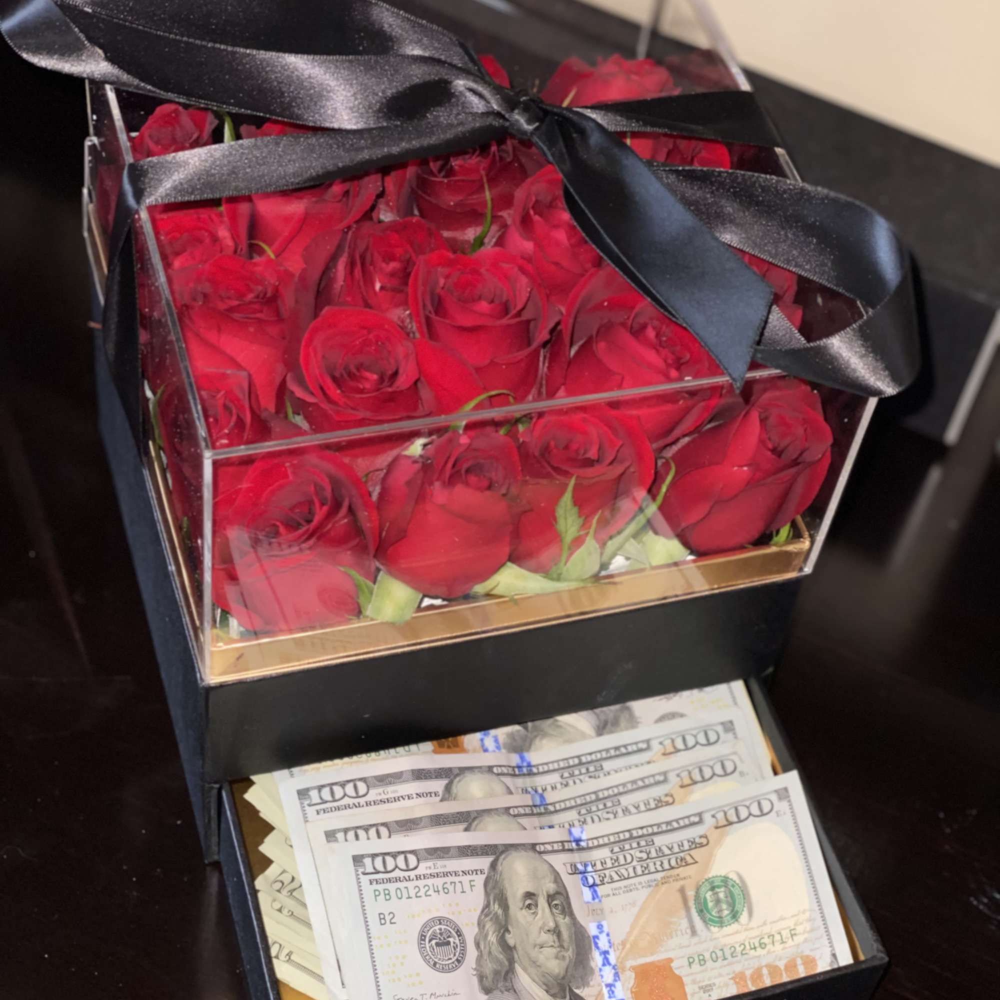 Red roses in a clear gift box with a black ribbon, above a drawer of cash