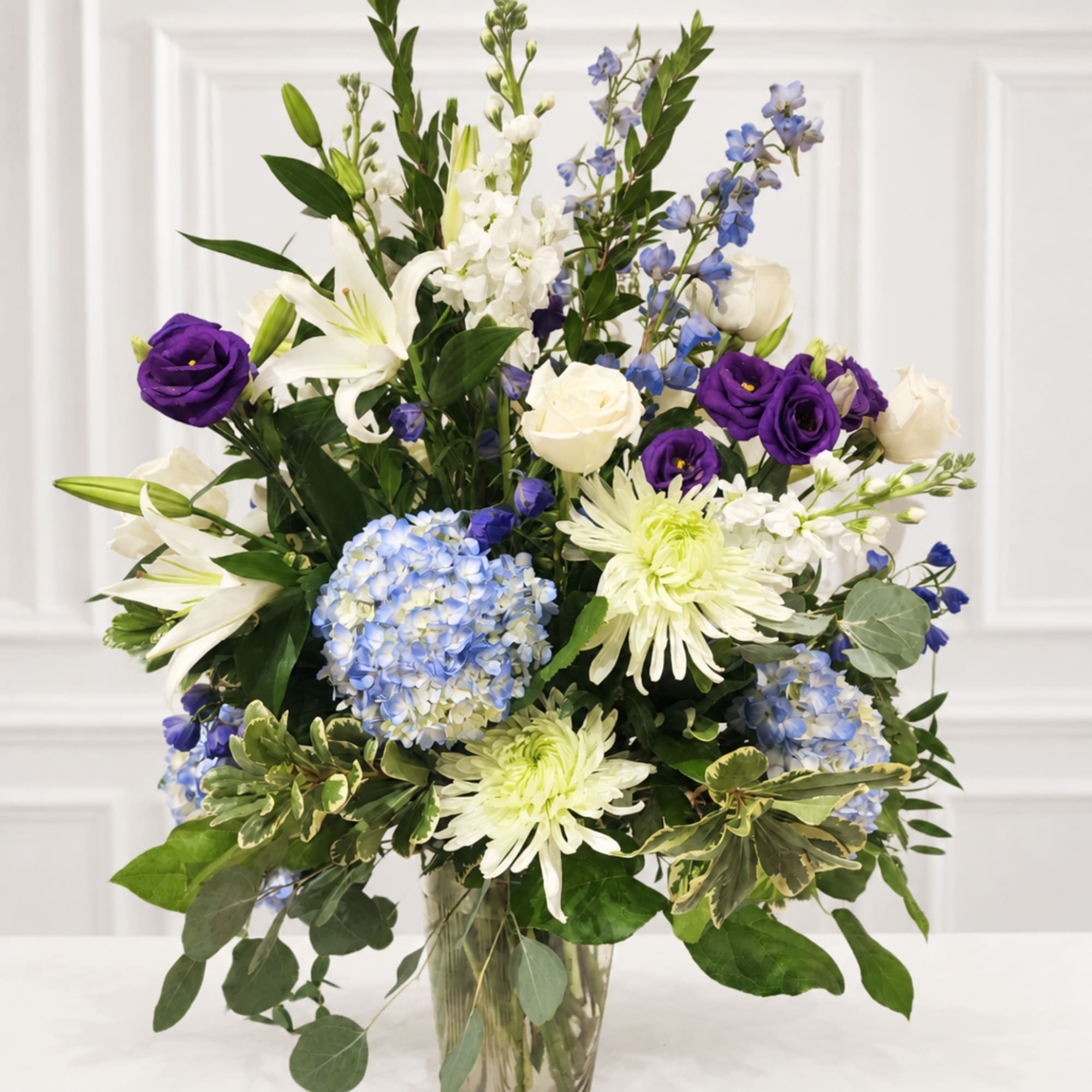 Tall bouquet of white lilies, purple roses, and blue flowers in a glass vase