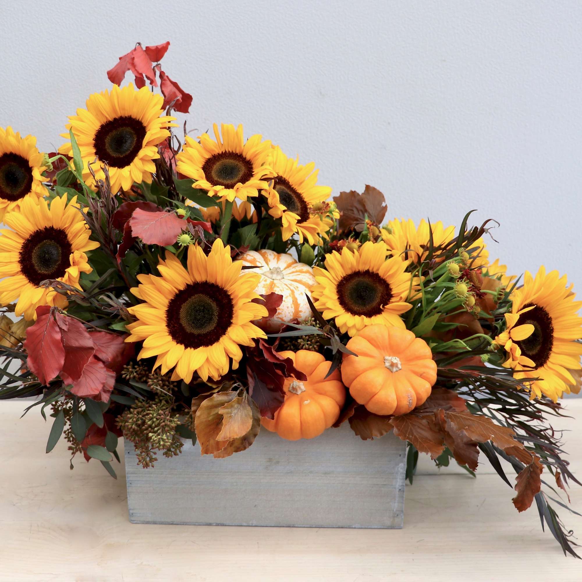 Low rectangular arrangement of yellow sunflowers with small orange pumpkins in a wooden box