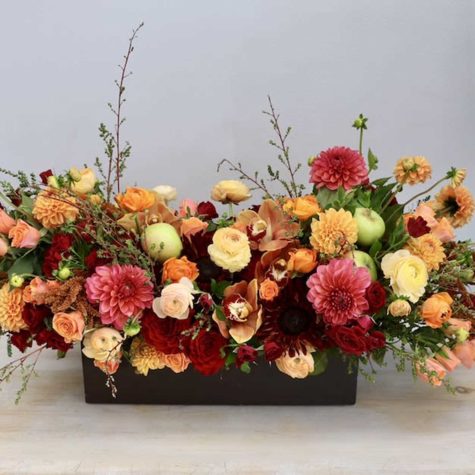 Low rectangular centerpiece of red, peach, and yellow blooms in a dark container