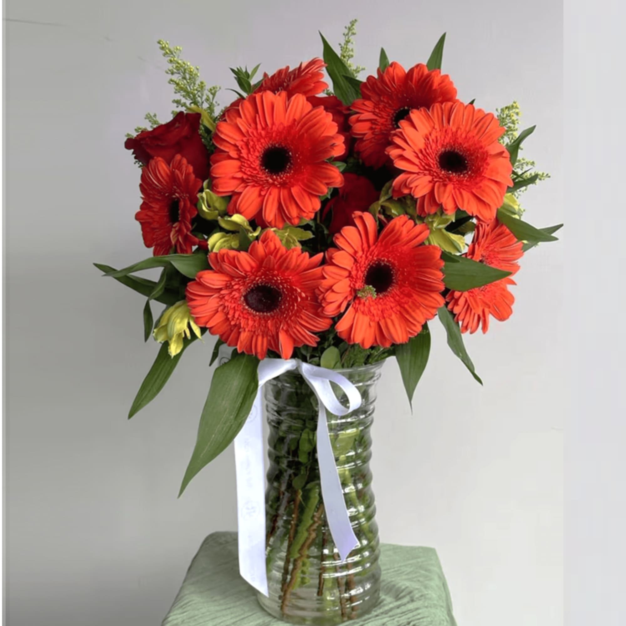 Tall glass vase of bright orange gerbera daisies with red roses and yellow accent blooms tied with a white ribbon.