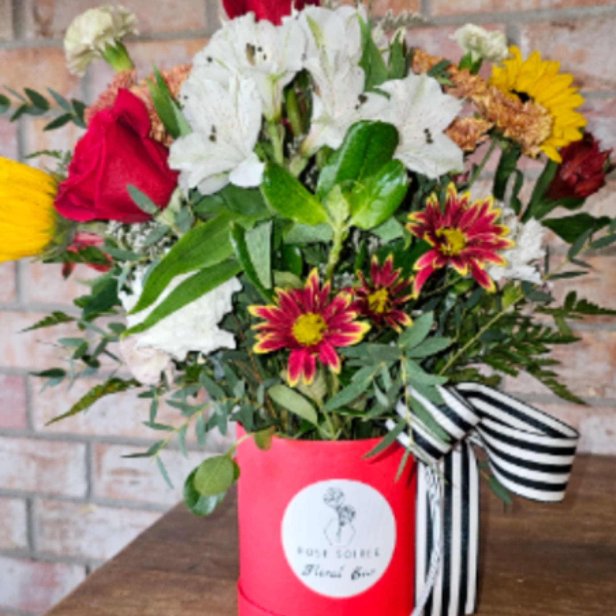 Mixed arrangement of red roses, white blooms, and yellow flowers in a red hatbox with striped ribbon