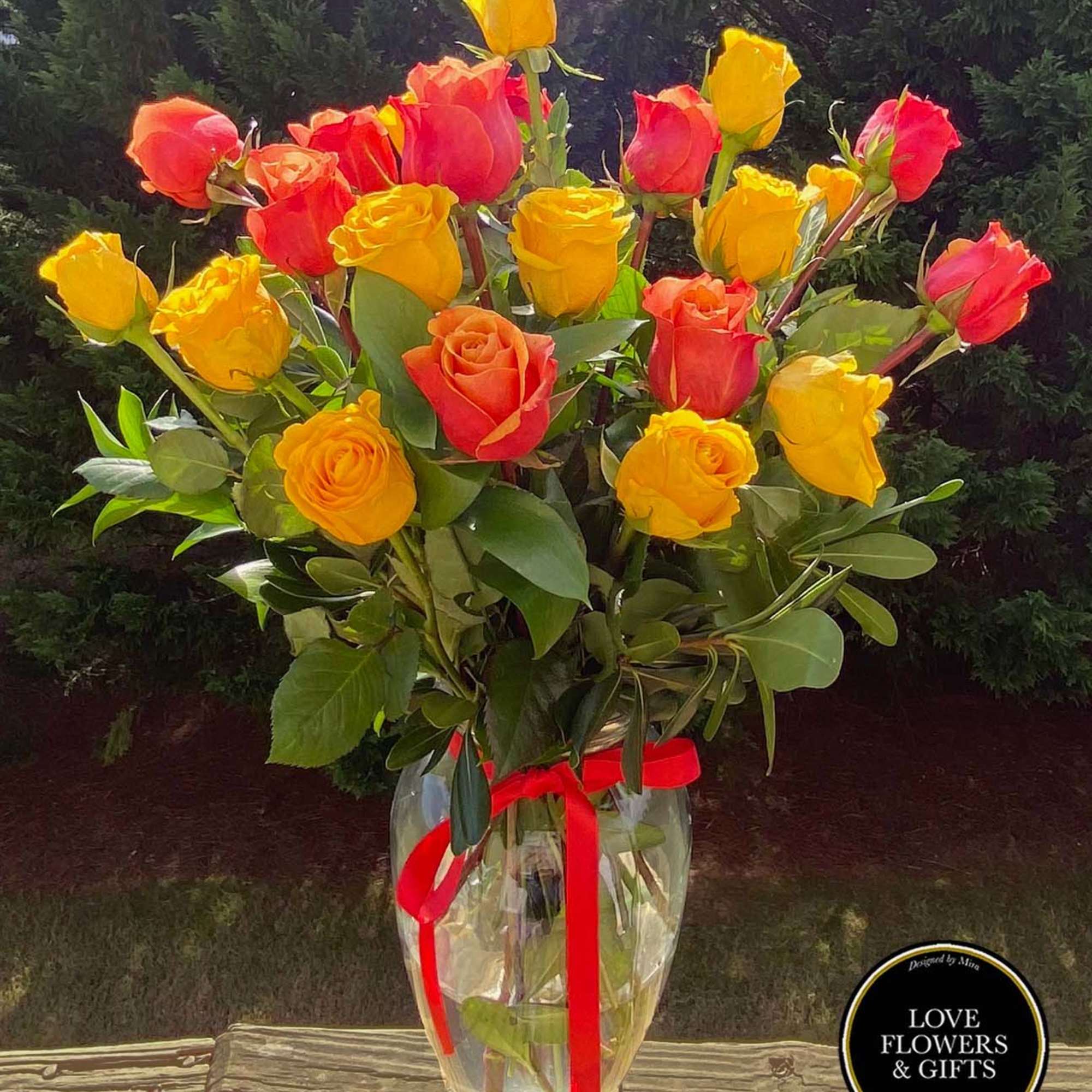 Tall bouquet of yellow and orange roses in a clear glass vase with red ribbon