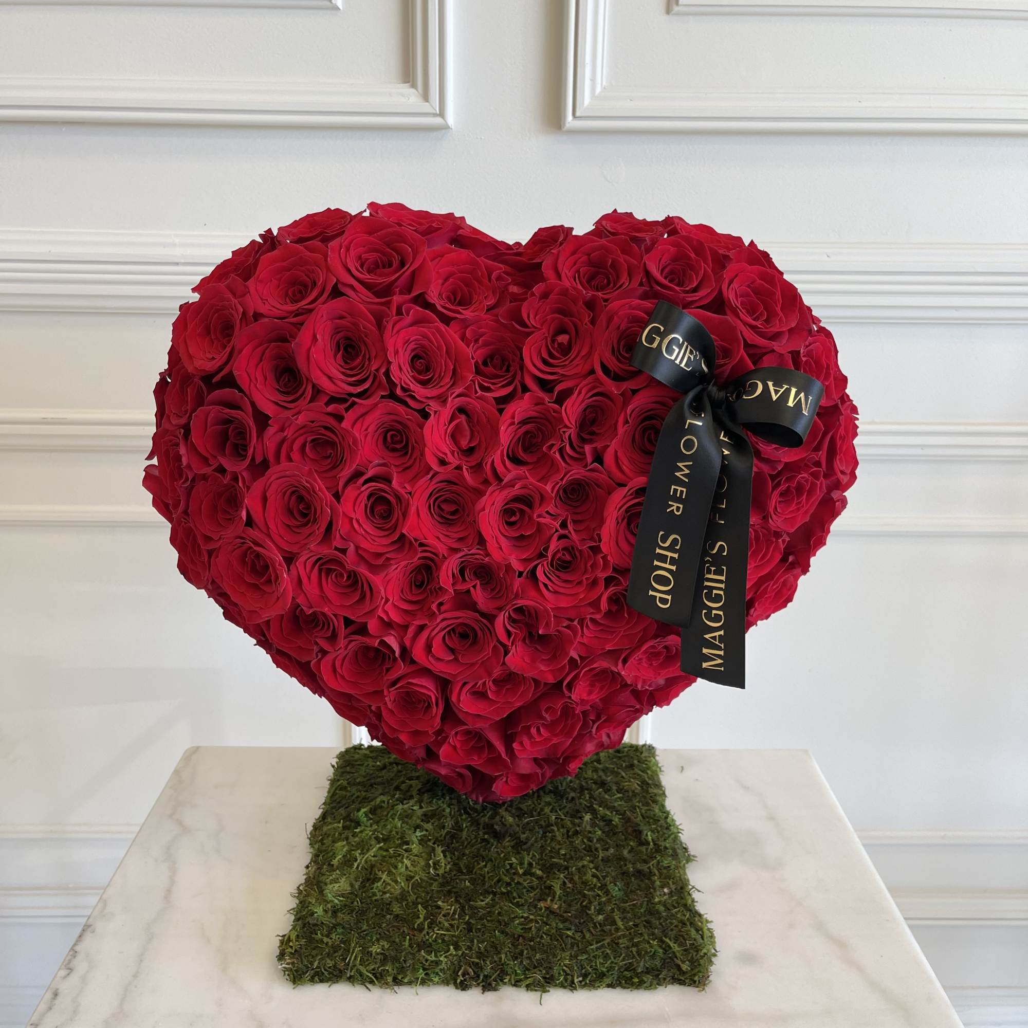 Heart-shaped arrangement of red roses on a moss-covered base with a black ribbon