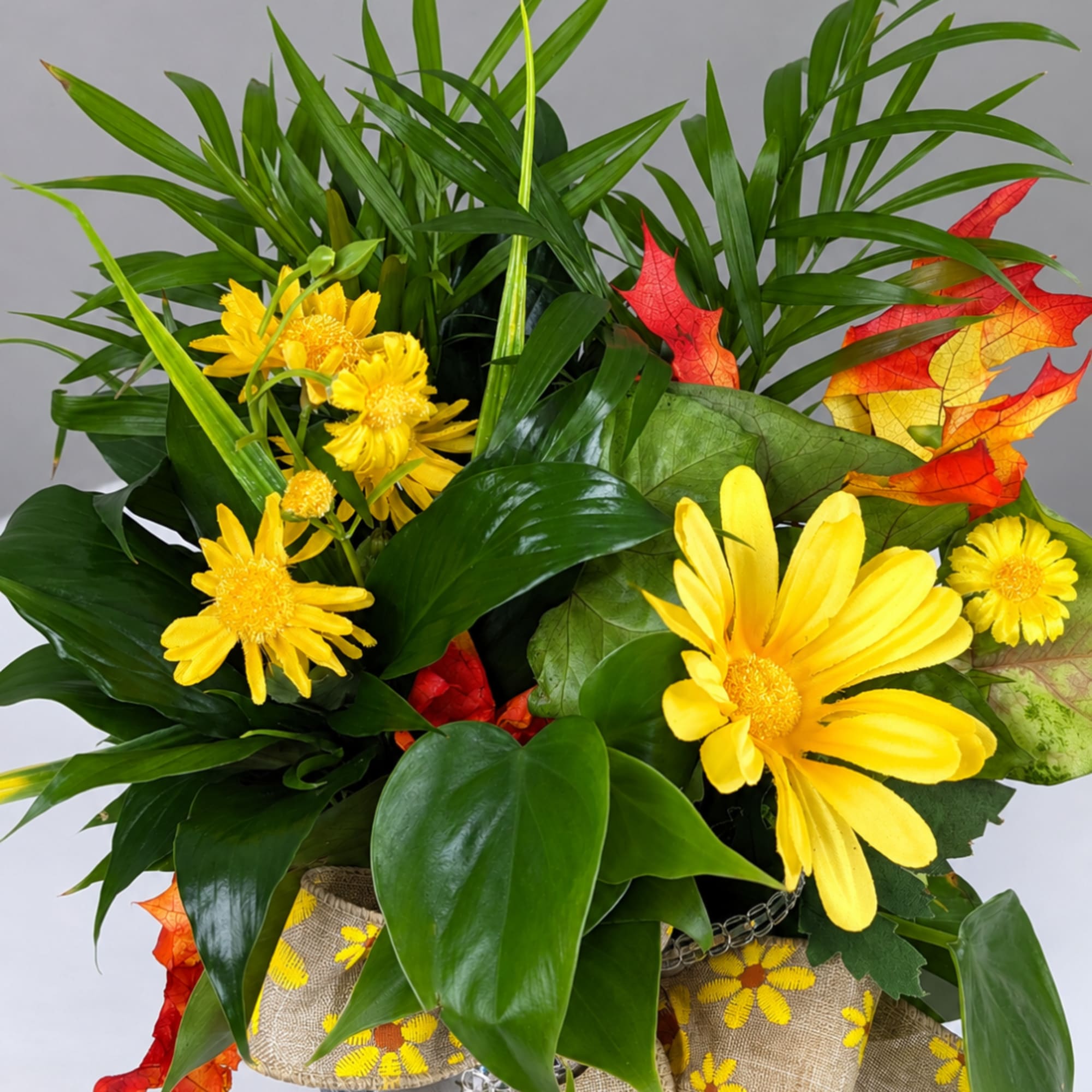 Yellow daisy bouquet with tropical leaves and autumn-colored accents in a decorative container