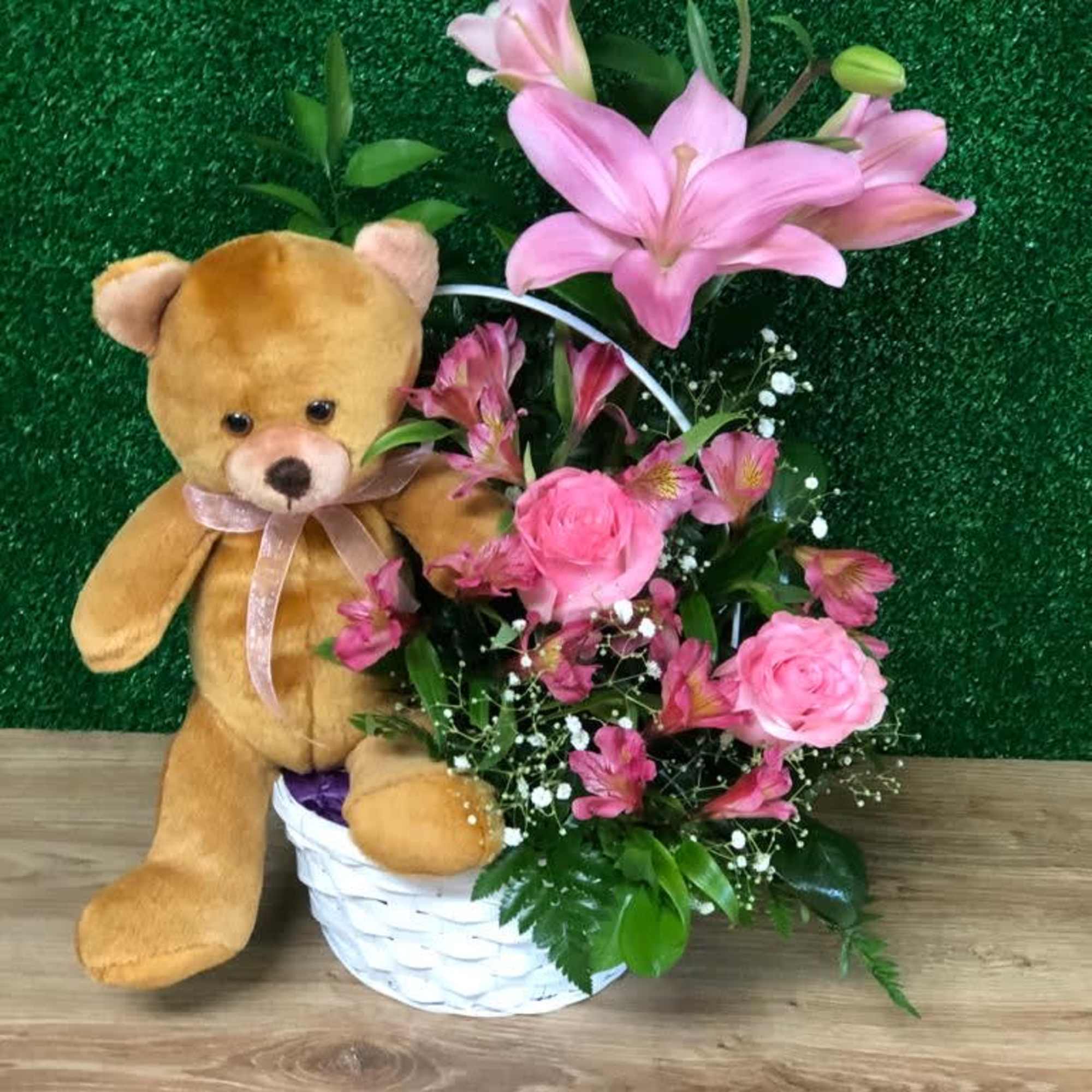 Tan teddy bear beside pink lilies and roses in a white woven basket.