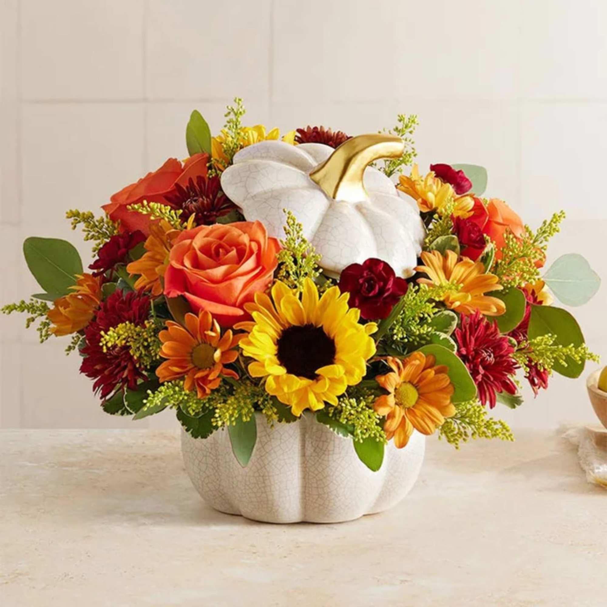 Autumn arrangement of sunflowers, orange roses, and mums in a white ceramic pumpkin container with gold stem.