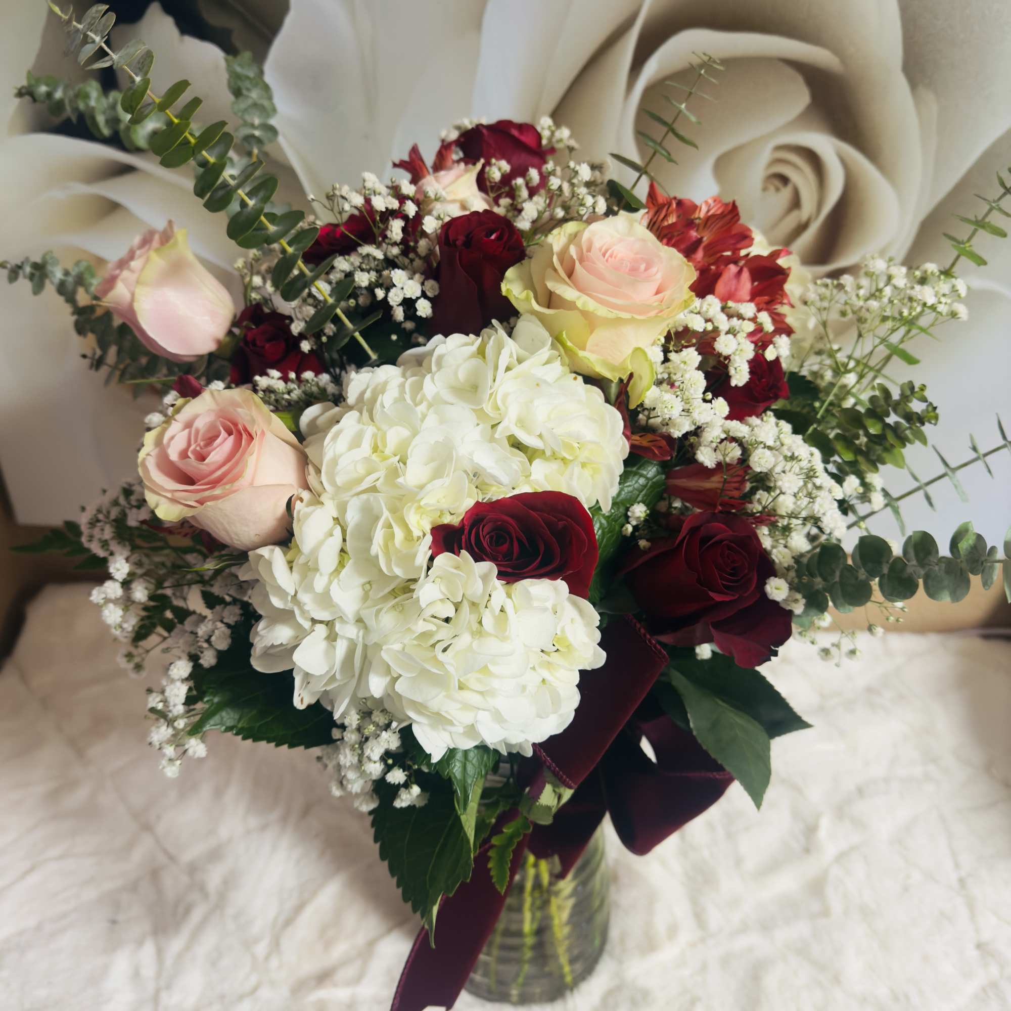 Vase arrangement of white hydrangeas with red and pink roses and baby's breath in a clear glass vase with burgundy ribbon