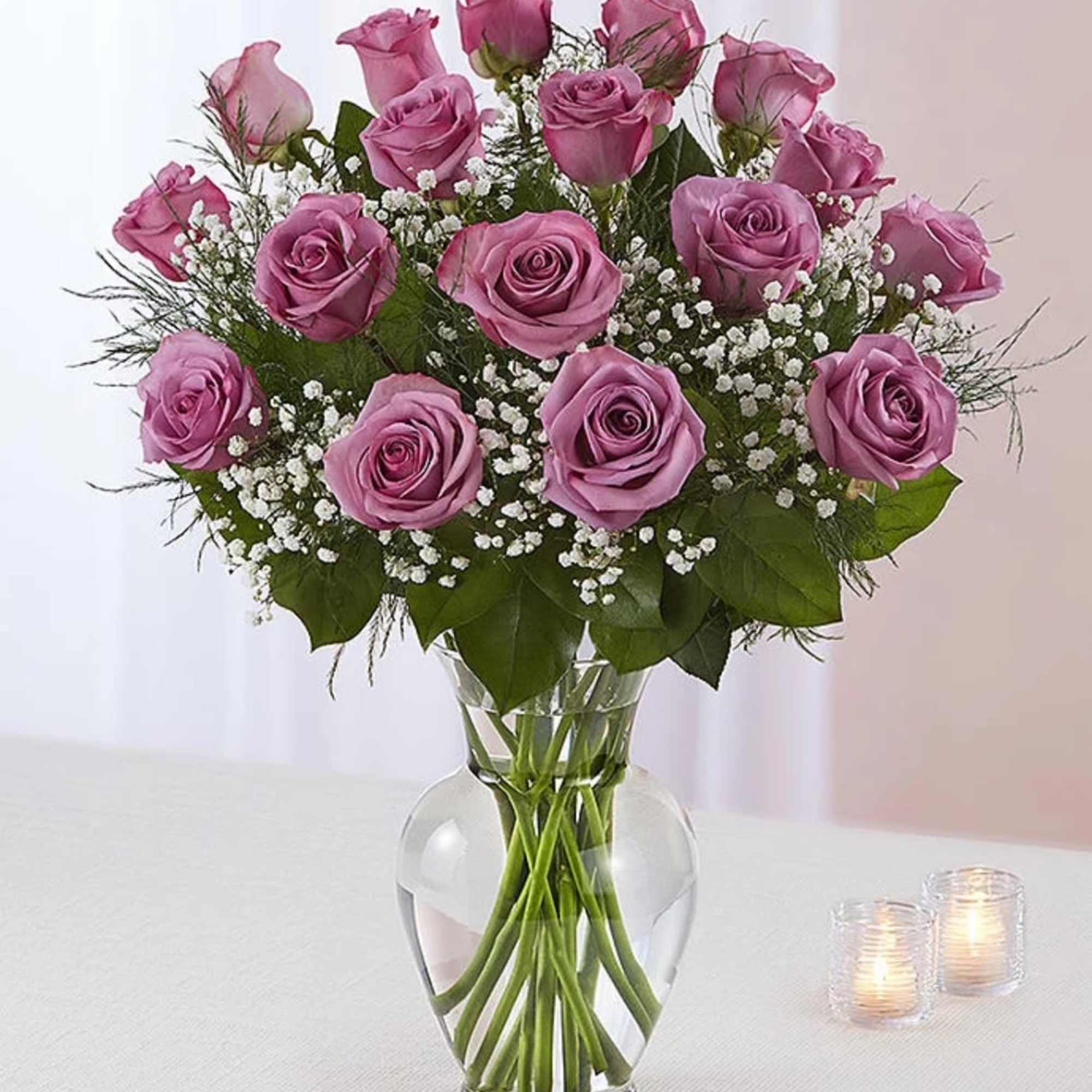 Glass vase of lavender roses with white filler flowers and greenery, with two small votive candles beside it