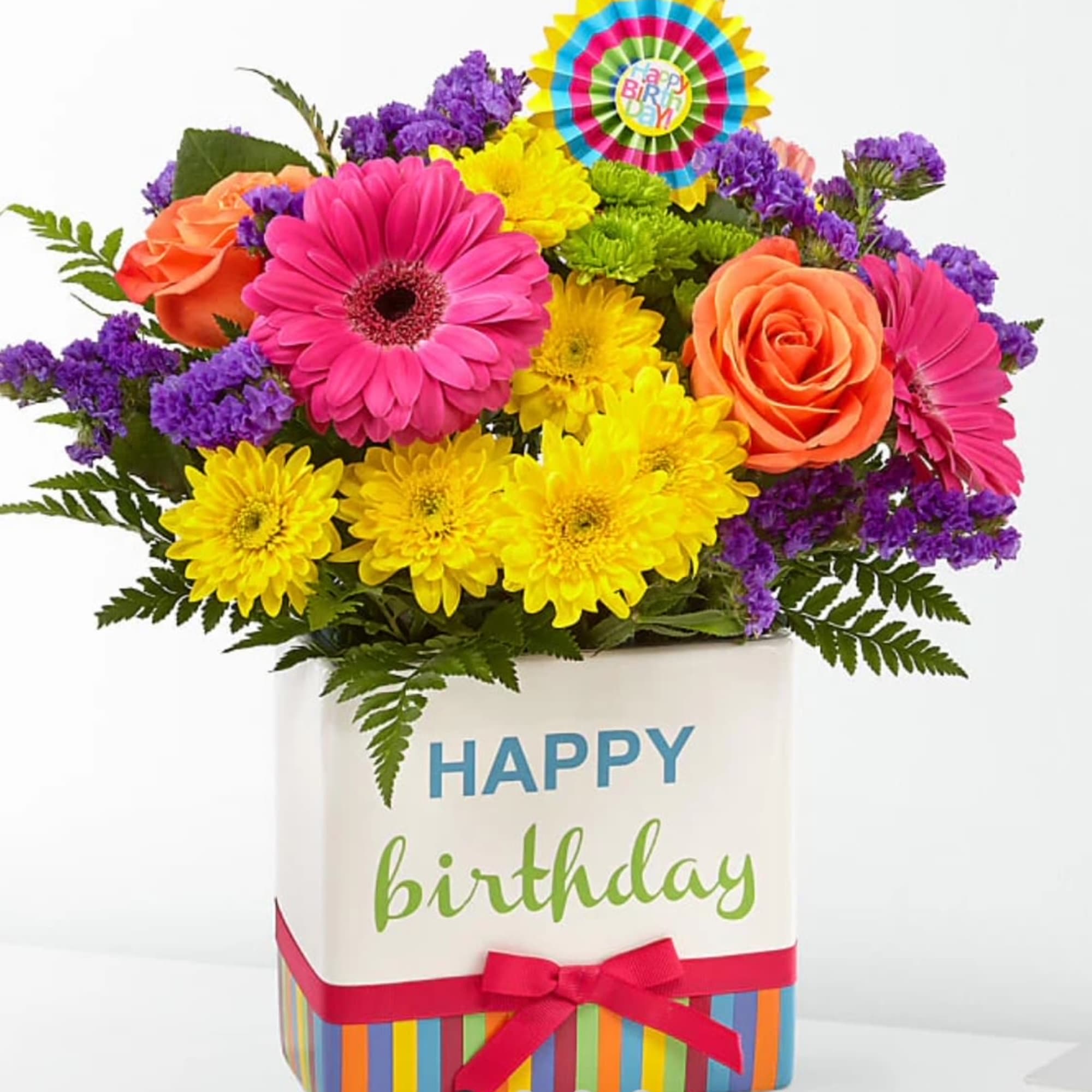 Bright birthday arrangement with pink gerbera daisies, orange roses, yellow mums in a Happy Birthday cube vase