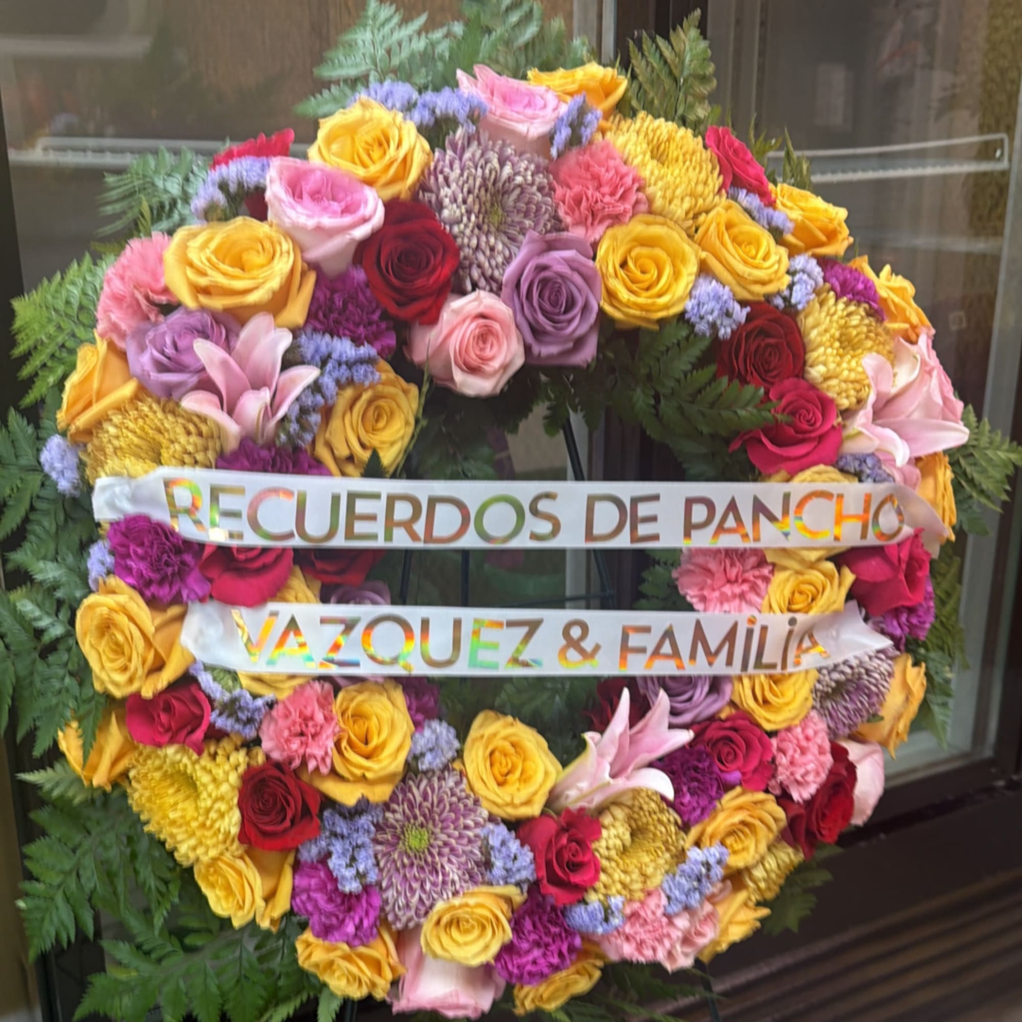 Round floral wreath with roses, lilies, and chrysanthemums and a memorial ribbon