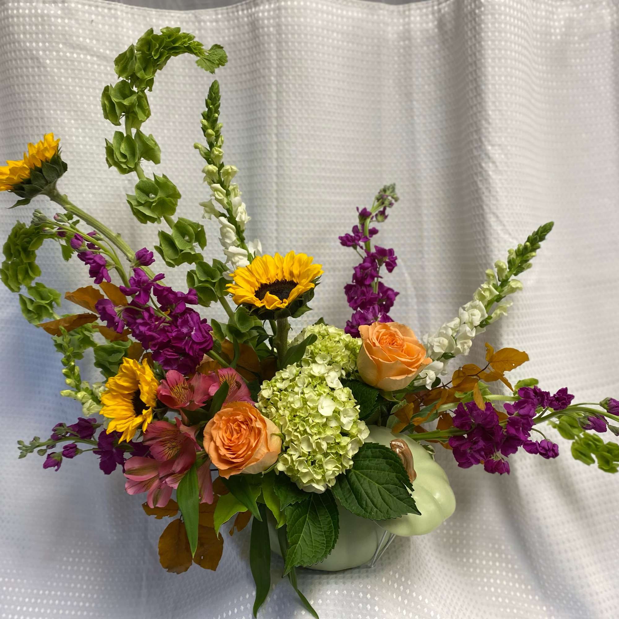 Mixed bouquet with sunflowers, peach roses, and purple blooms in a pale green pumpkin vase