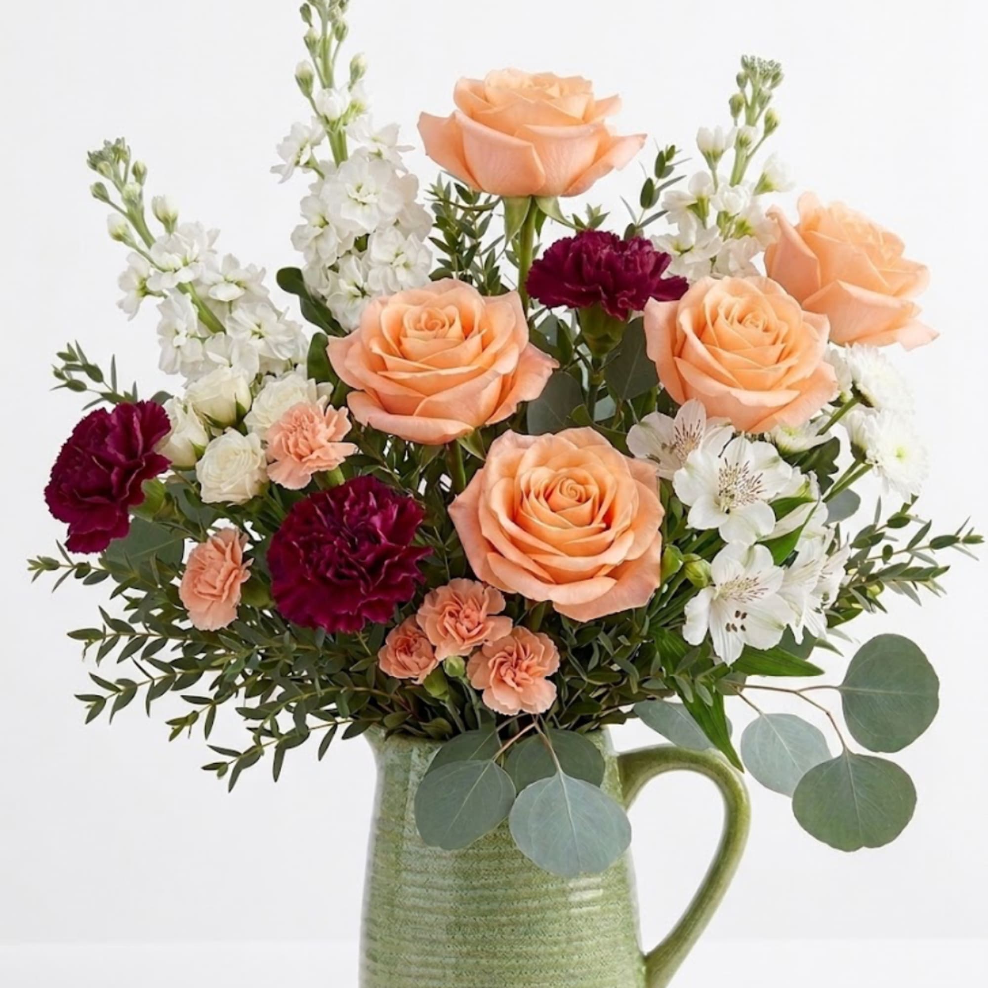 Peach roses, white flowers, and burgundy carnations in a green ceramic pitcher