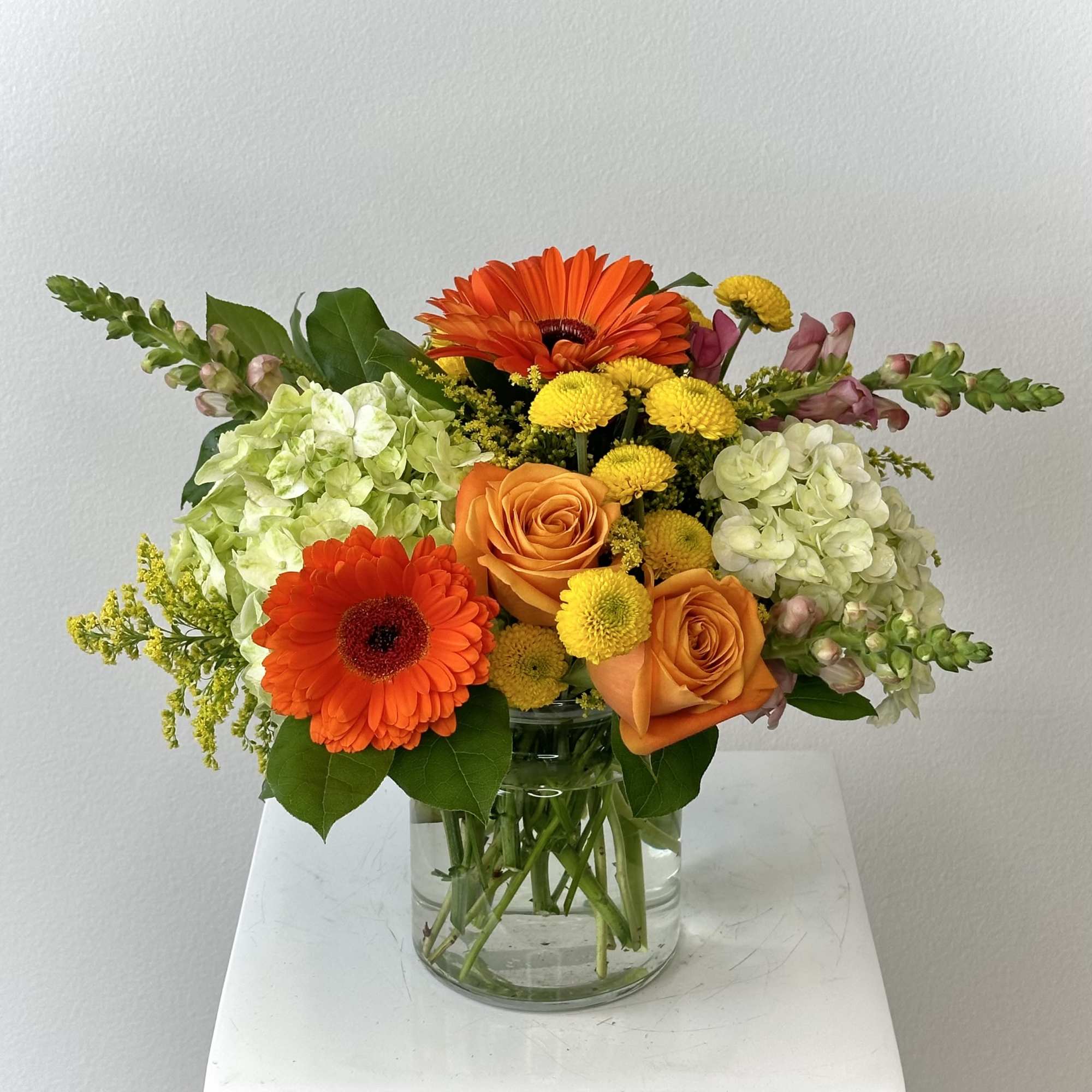 Orange gerbera daisies and roses in a clear glass vase with pale hydrangeas