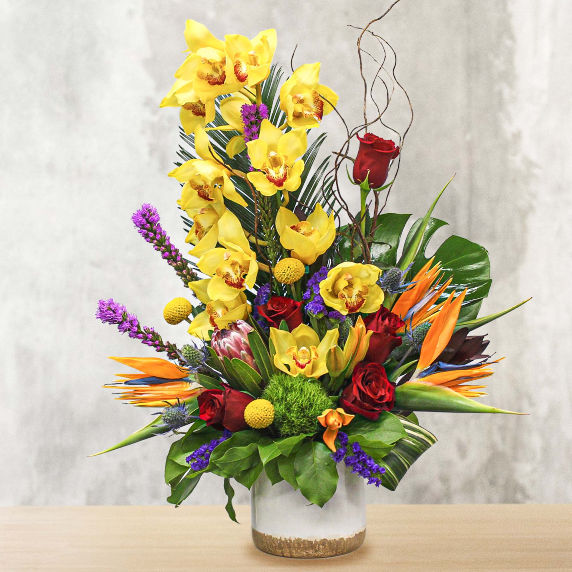 Tall tropical bouquet with yellow orchids, red roses, and orange bird-of-paradise in a glass vase