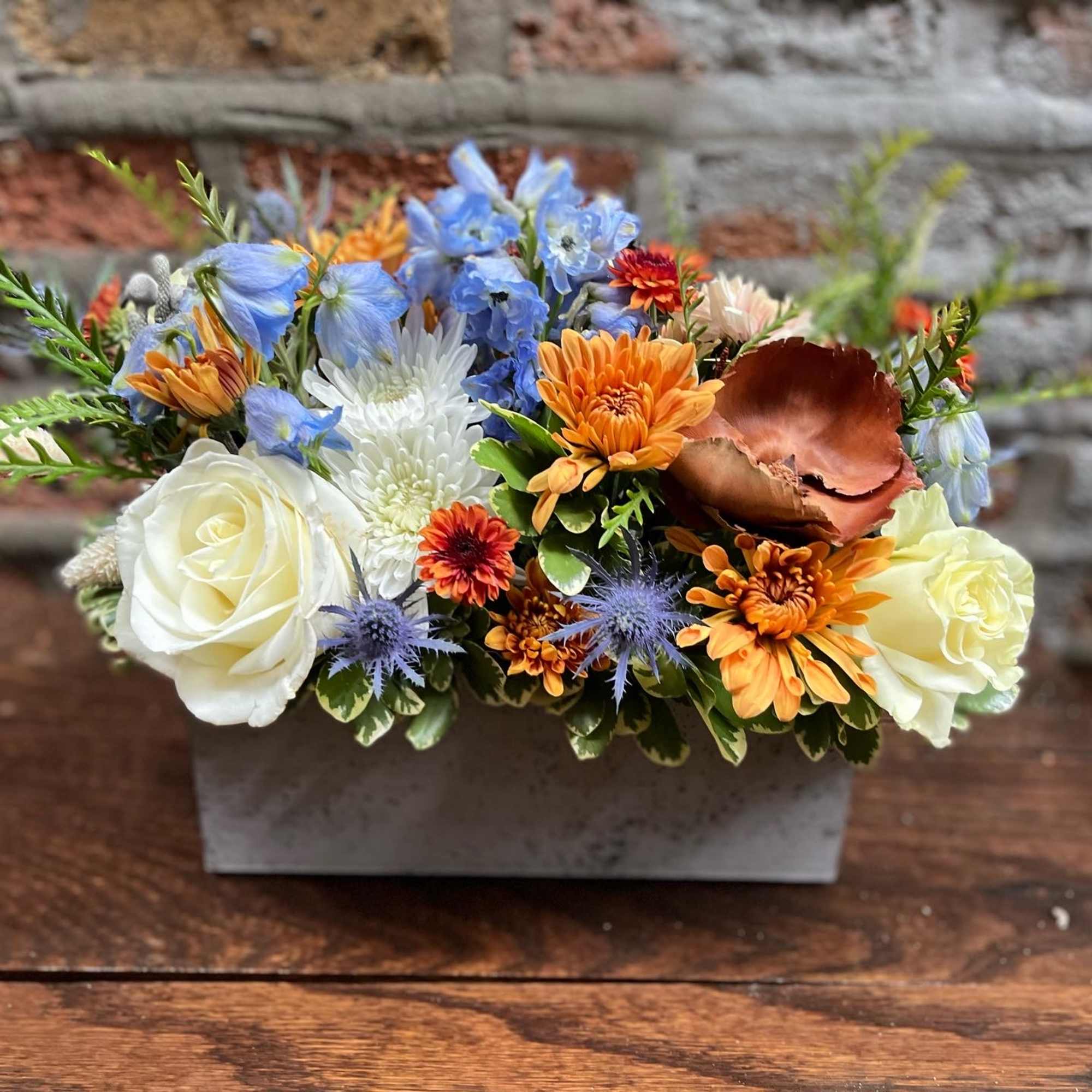 Mixed bouquet with white roses, blue flowers, and orange blooms in a gray container