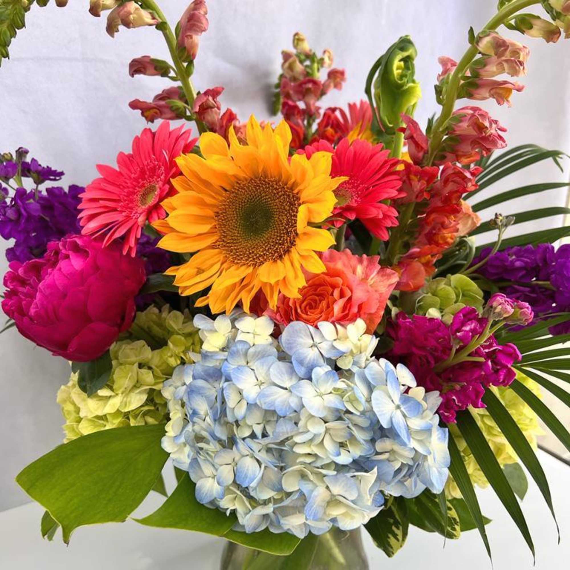 Tall mixed arrangement with sunflower, gerbera daisies, roses and hydrangeas in a clear glass vase